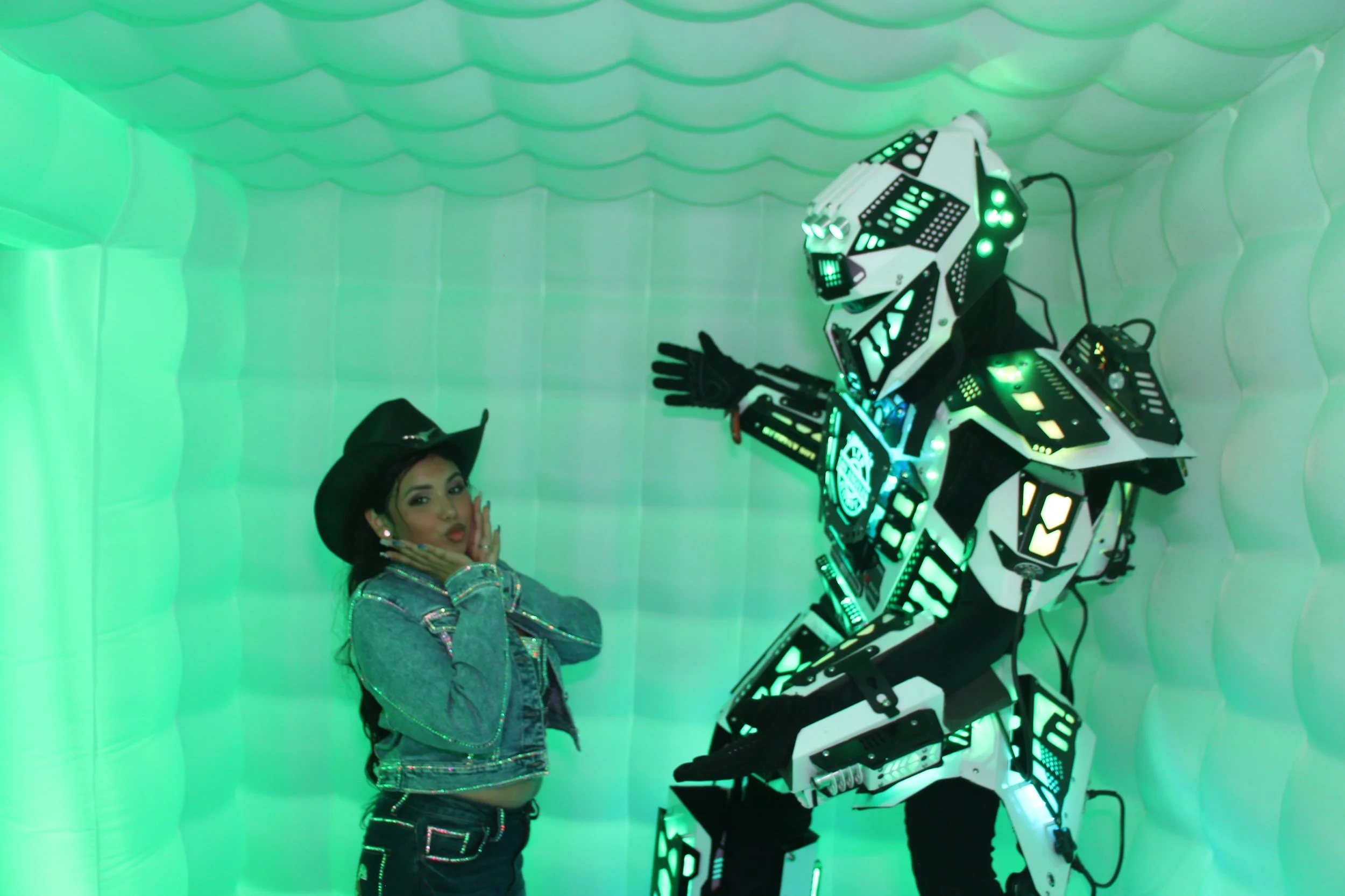 A woman in a cowboy hat and denim jacket poses with a futuristic humanoid robot in a green-lit, textured room.