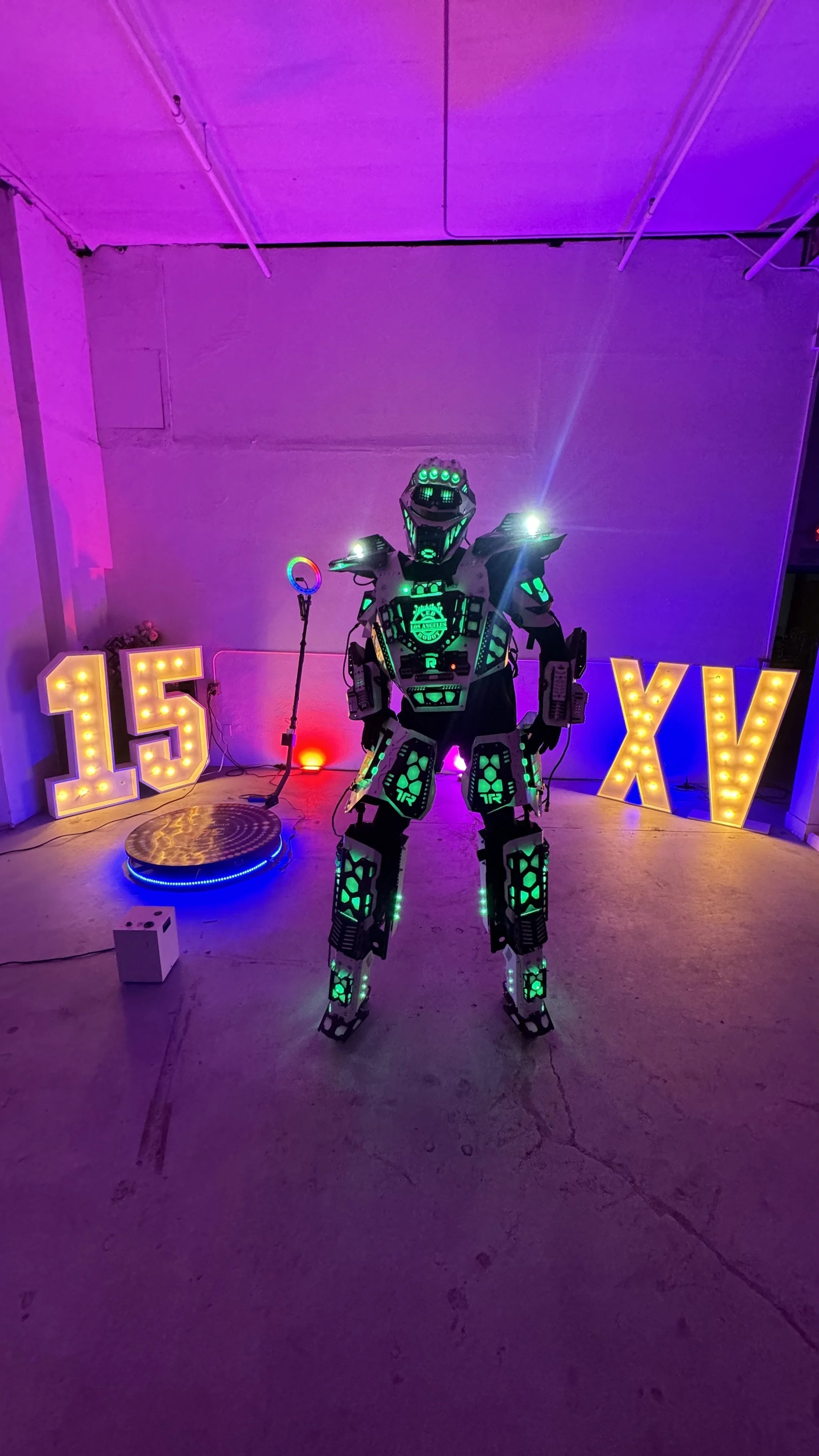A person in a futuristic, glowing robot costume at a neon birthday celebration with illuminated number letters on each side reading '15' and 'XV' in the background.