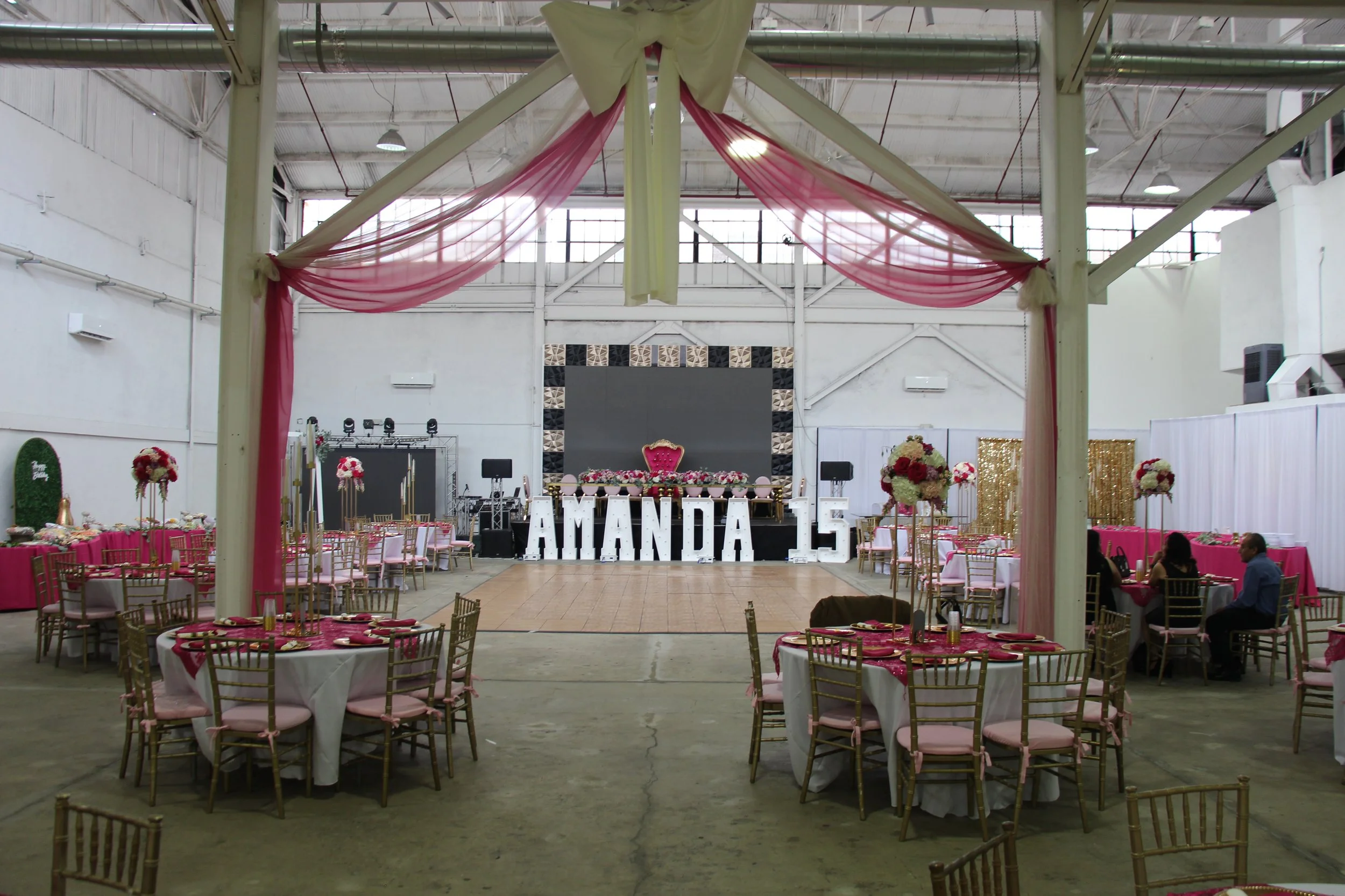 Event hall decorated for a birthday party with large white letter balloons spelling 'AMANDA 15' on the stage, pink and gold-themed tables, floral centerpieces, and a stage with a large screen and pink throne chair.