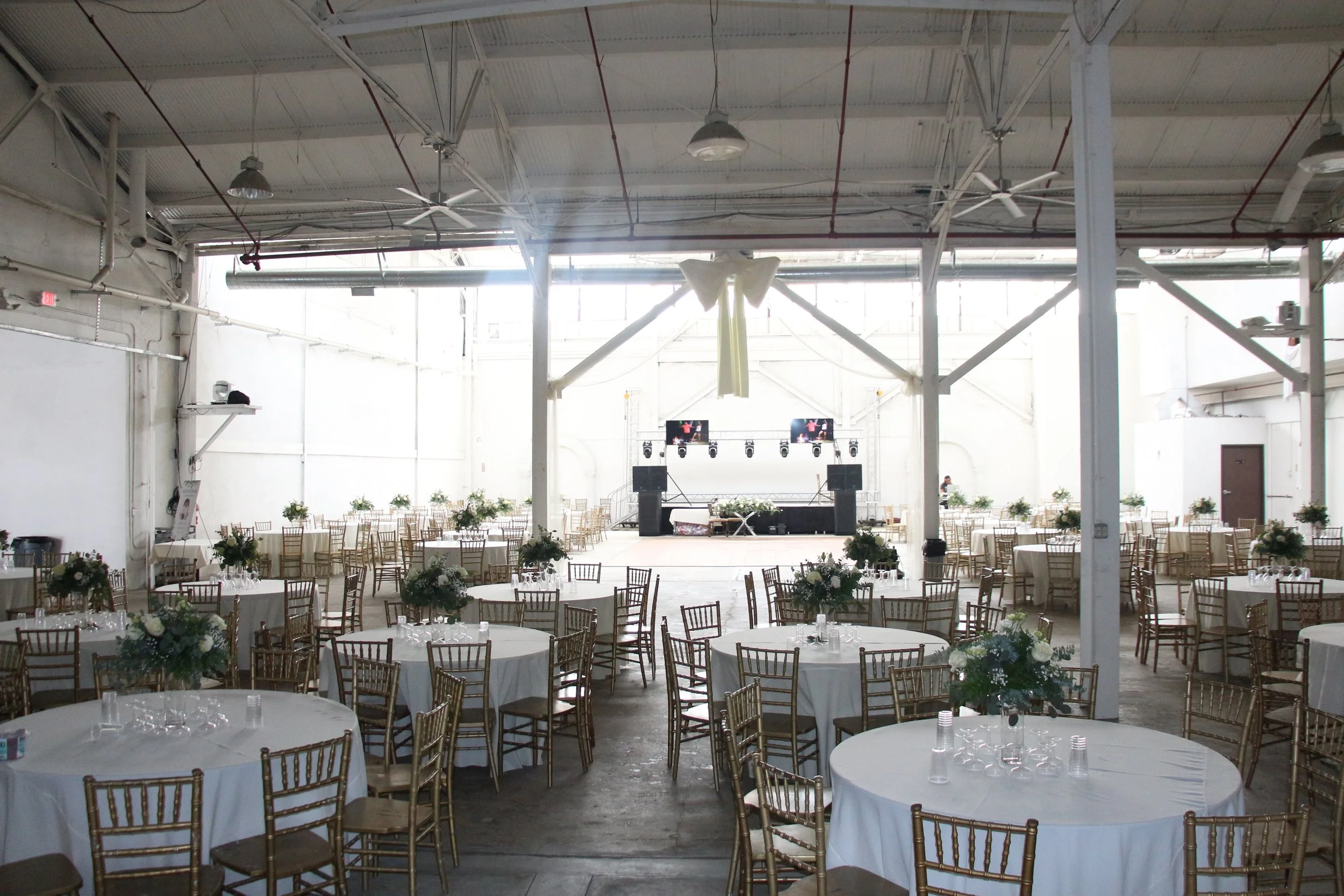 Interior of an event venue with round tables covered in white tablecloths, decorated with floral centerpieces, and surrounded by gold-colored chairs. A stage with lighting and sound equipment is set up at the far end, with bright natural light streaming in from large windows and an open ceiling with ceiling fans.