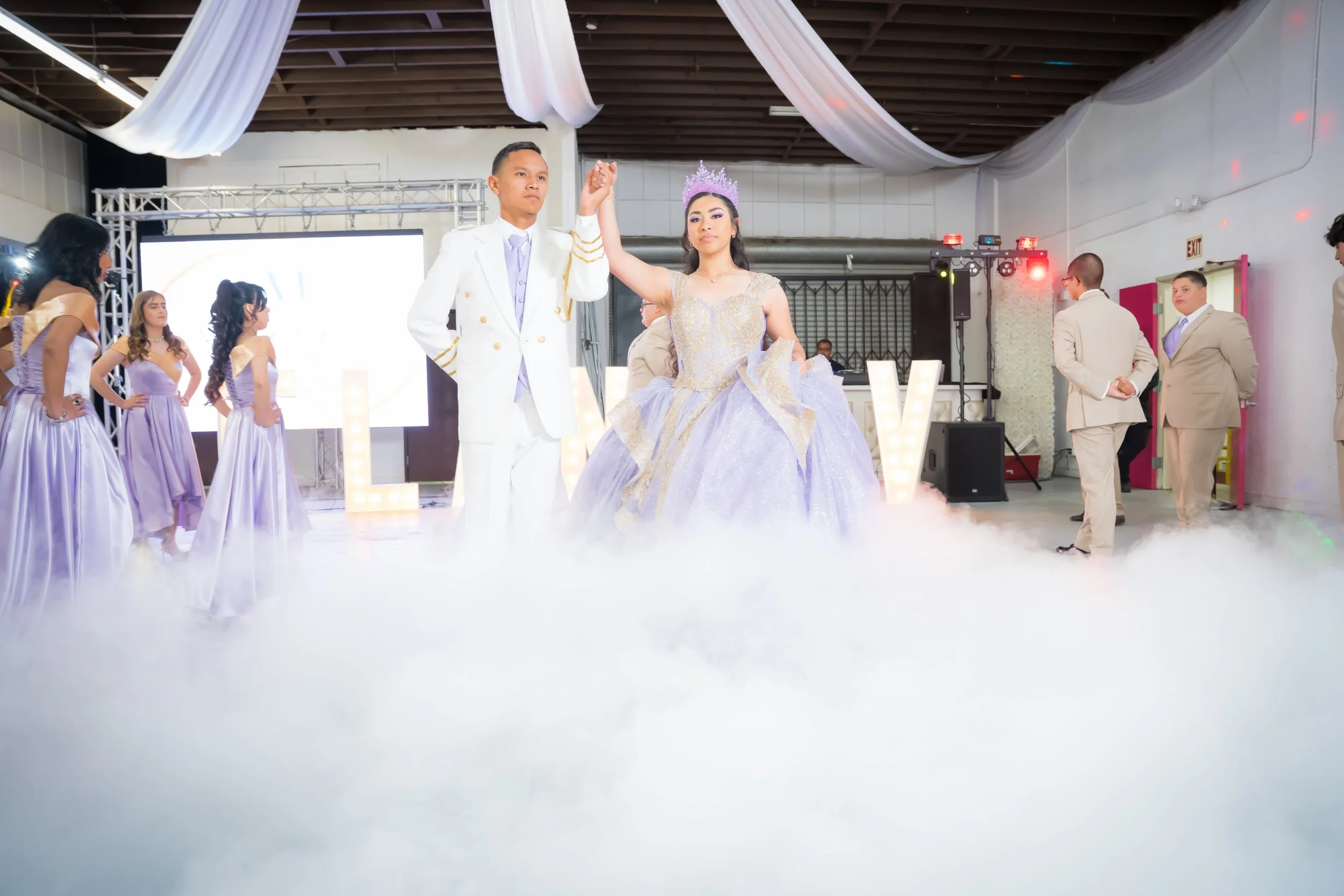 A couple dances at a wedding reception, with a bride wearing a lavender gown and a crown, and a groom in a white suit, surrounded by guests and a fog effect.