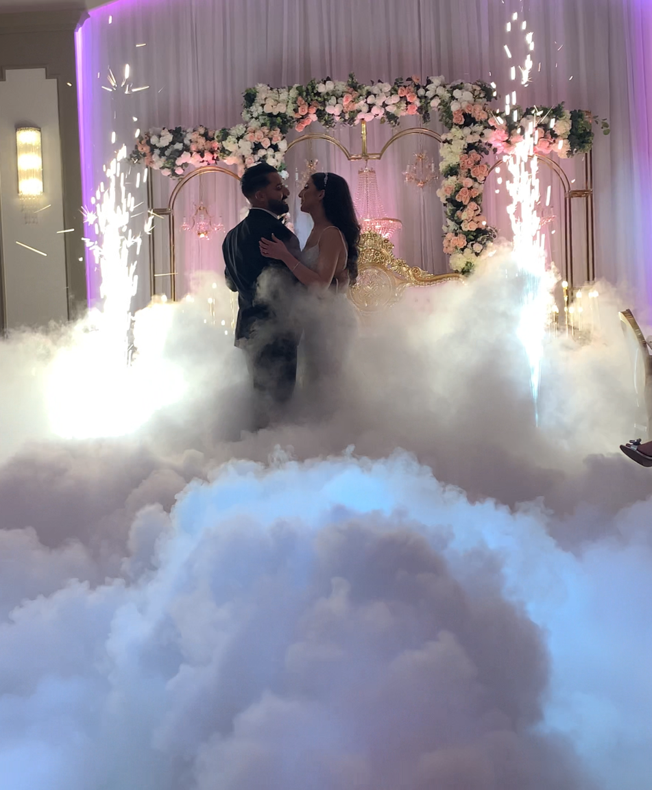 A couple sharing a dance at their wedding reception, surrounded by fog, sparkler fireworks, and floral decorations on a decorated stage.