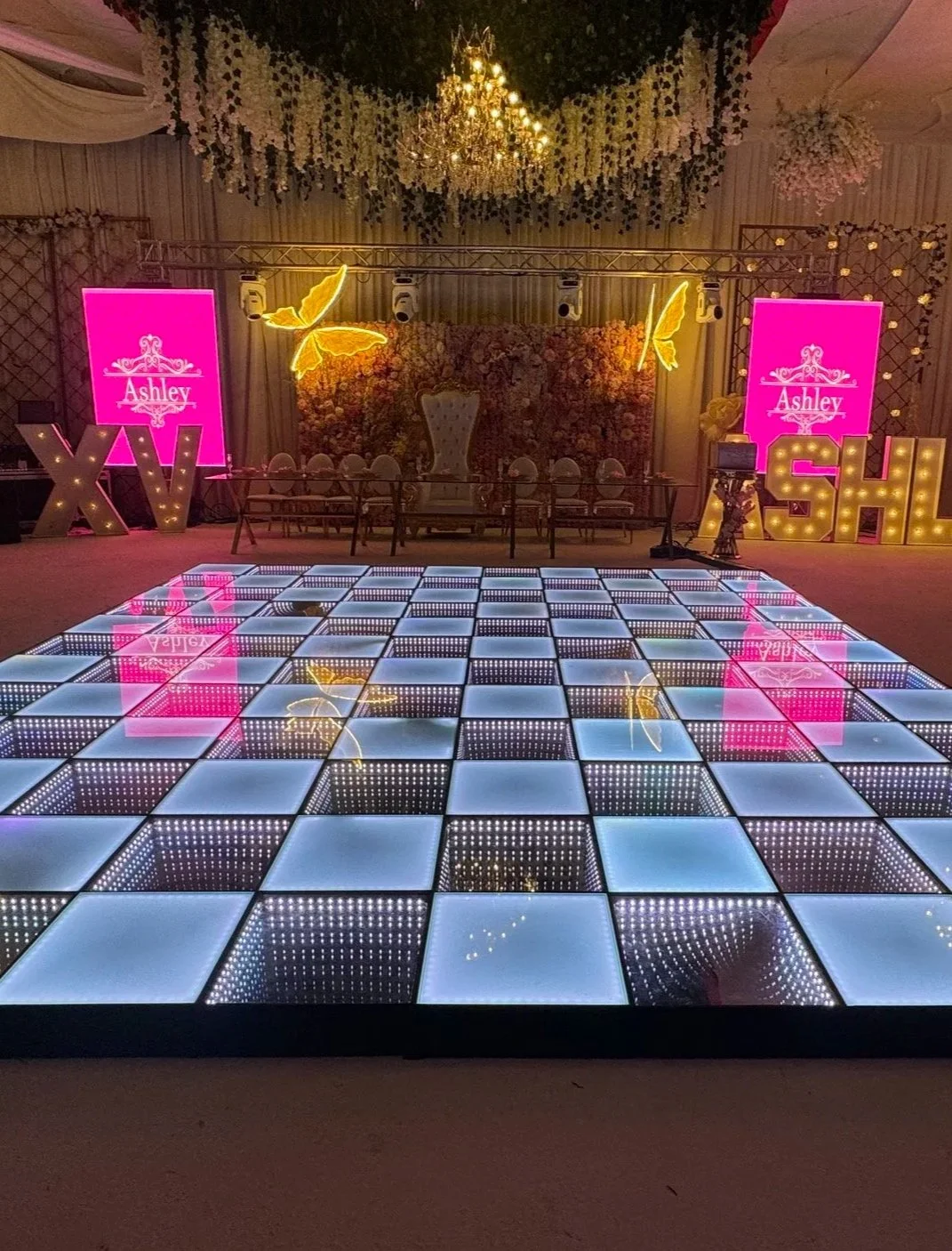 A decorated event space with a lit-up dance floor, floral backdrop, large illuminated letters spelling 'XV', and pink screens with the name Ashley. There are large butterfly decorations and overhead chandeliers.
