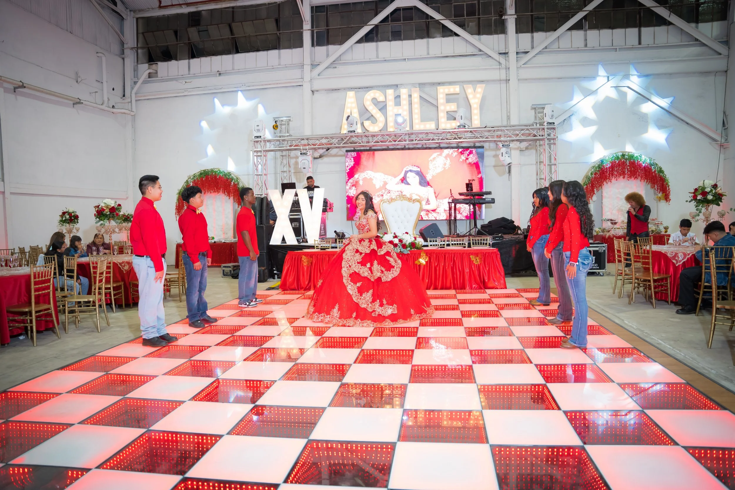 A birthday celebration with a young woman in a red and gold dress sitting on a throne-like chair in the center of a dance floor. Six people, three boys and three girls, stand facing her in a line on either side of the dance floor. The background feat