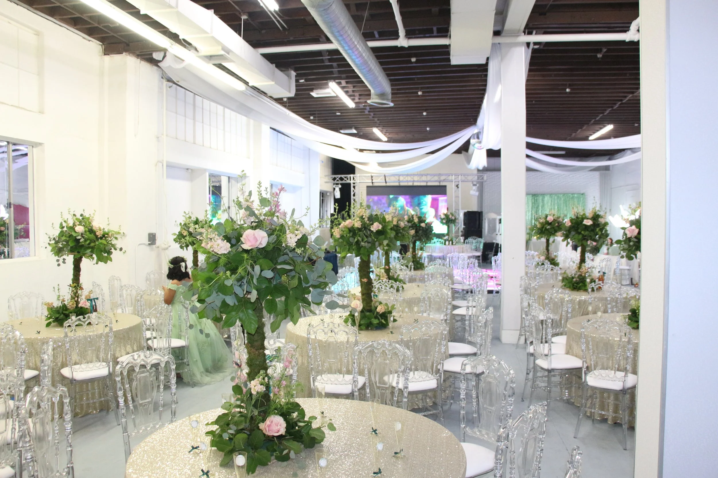 Event space decorated with floral centerpieces, round tables with glittery gold tablecloths, clear ghost chairs, white draping on the ceiling, and a large screen at the back.