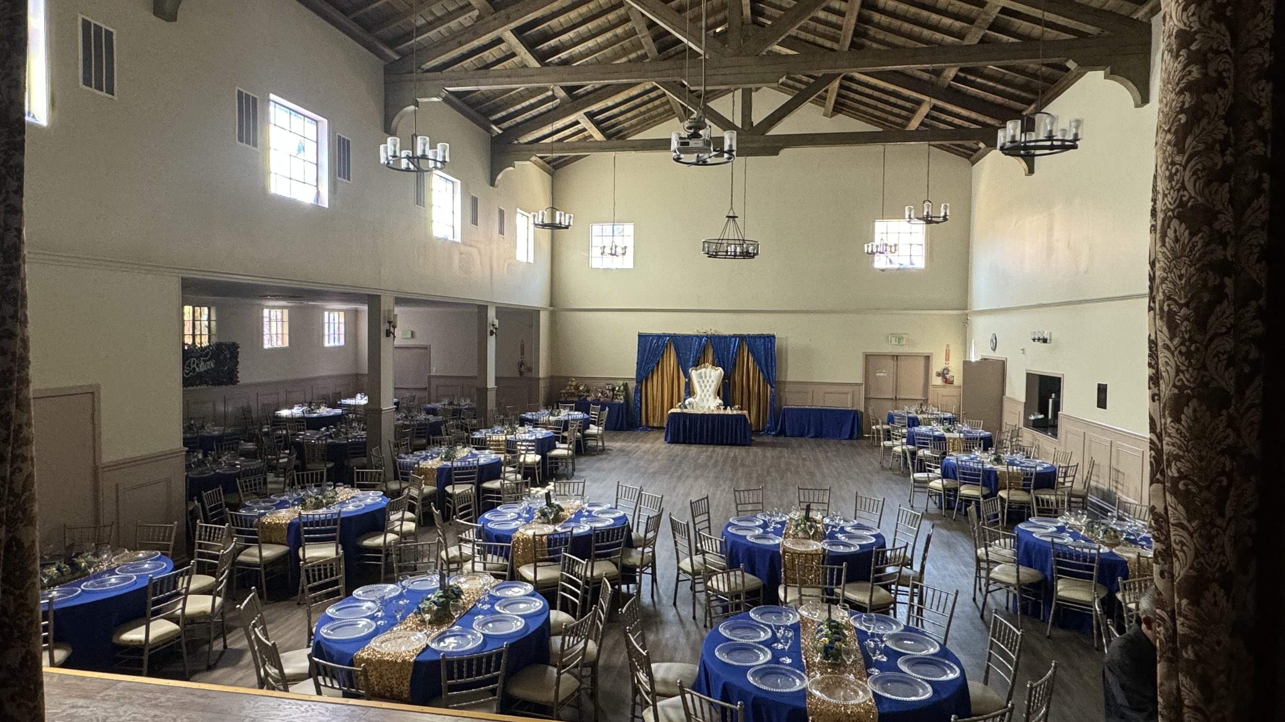 An event hall set up for a celebration with round tables decorated with blue tablecloths and gold runners, arranged around a large wooden dance floor, with a stage at the far end featuring a white throne-like chair, and decorated with blue and gold c