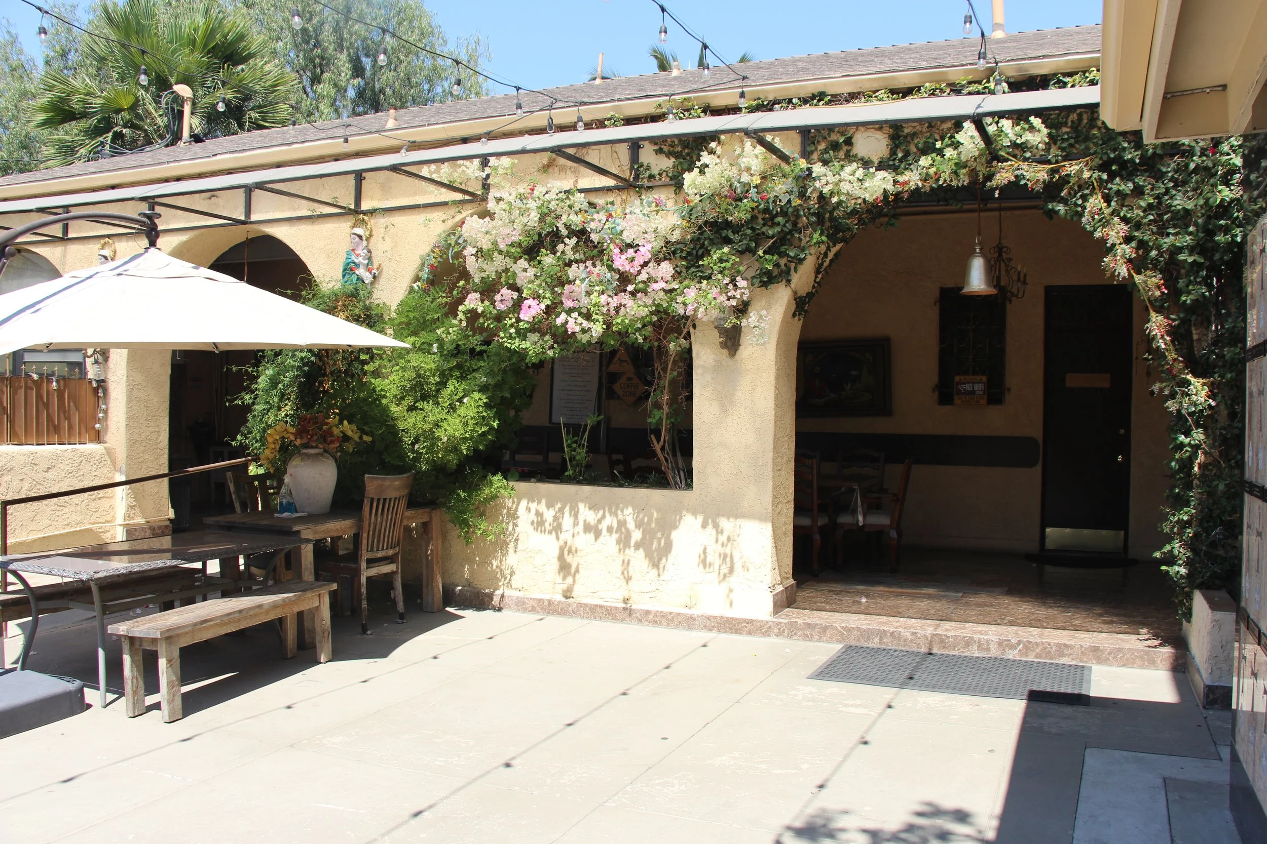 An outdoor patio of a restaurant with a large white umbrella, wooden benches, and a table with a vase of flowers. The building has archways, hanging string lights, and green and pink flowering vines.
