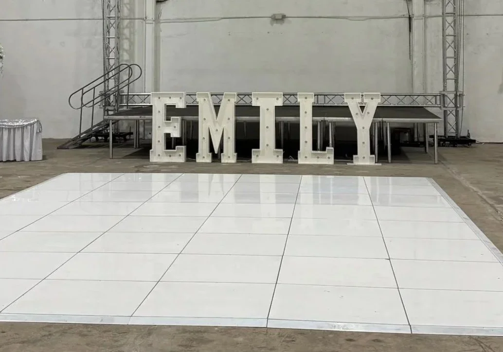Large white illuminated letters spelling out 'EMILY' placed on a stage with a metal platform, adjacent to a white dance floor in a spacious indoor event venue.