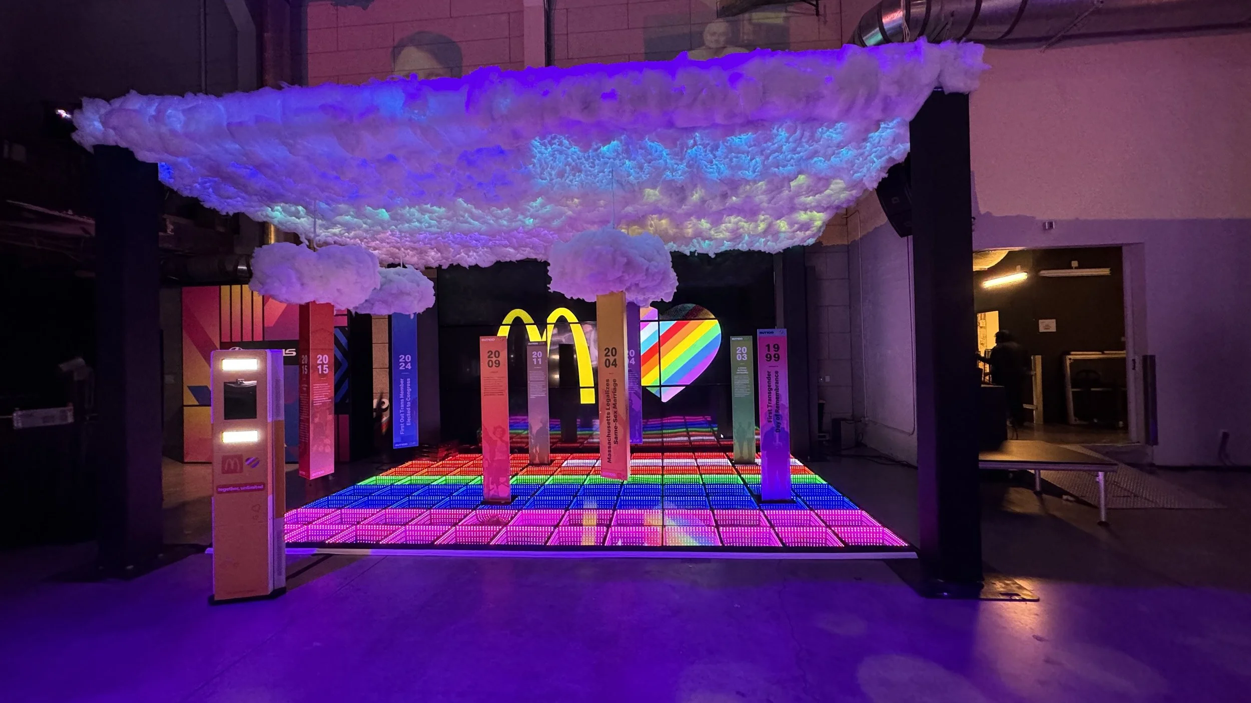 Colorful installation featuring neon rainbow heart and McDonald's logo, with rainbow-themed vertical banners and a cloud-like ceiling decoration in a dark room.