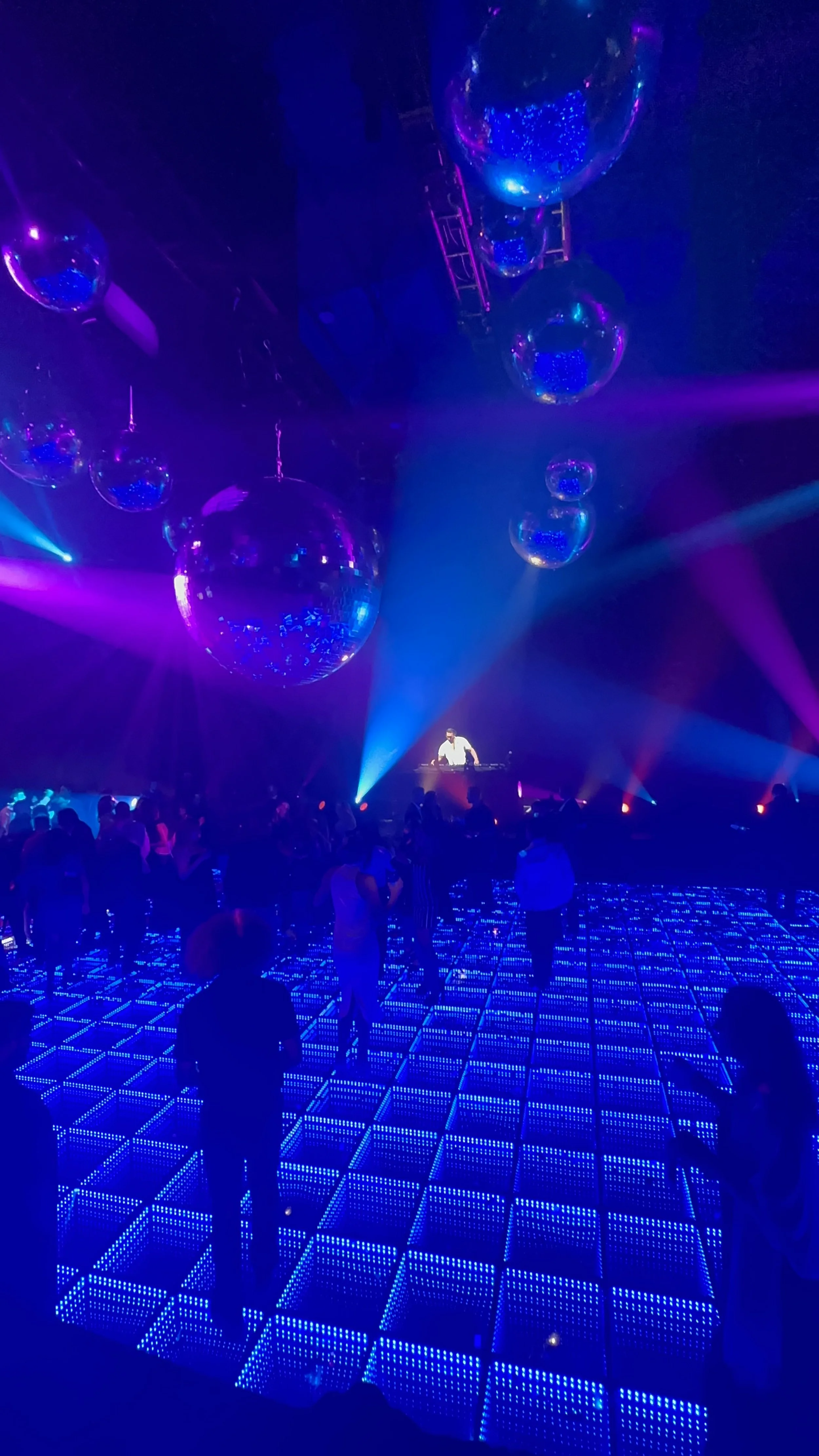 A lively nightclub scene with a DJ on stage and large disco balls hanging from the ceiling. The dance floor is illuminated with blue LED lights, and people are dancing and socializing under colorful purple and blue lighting.