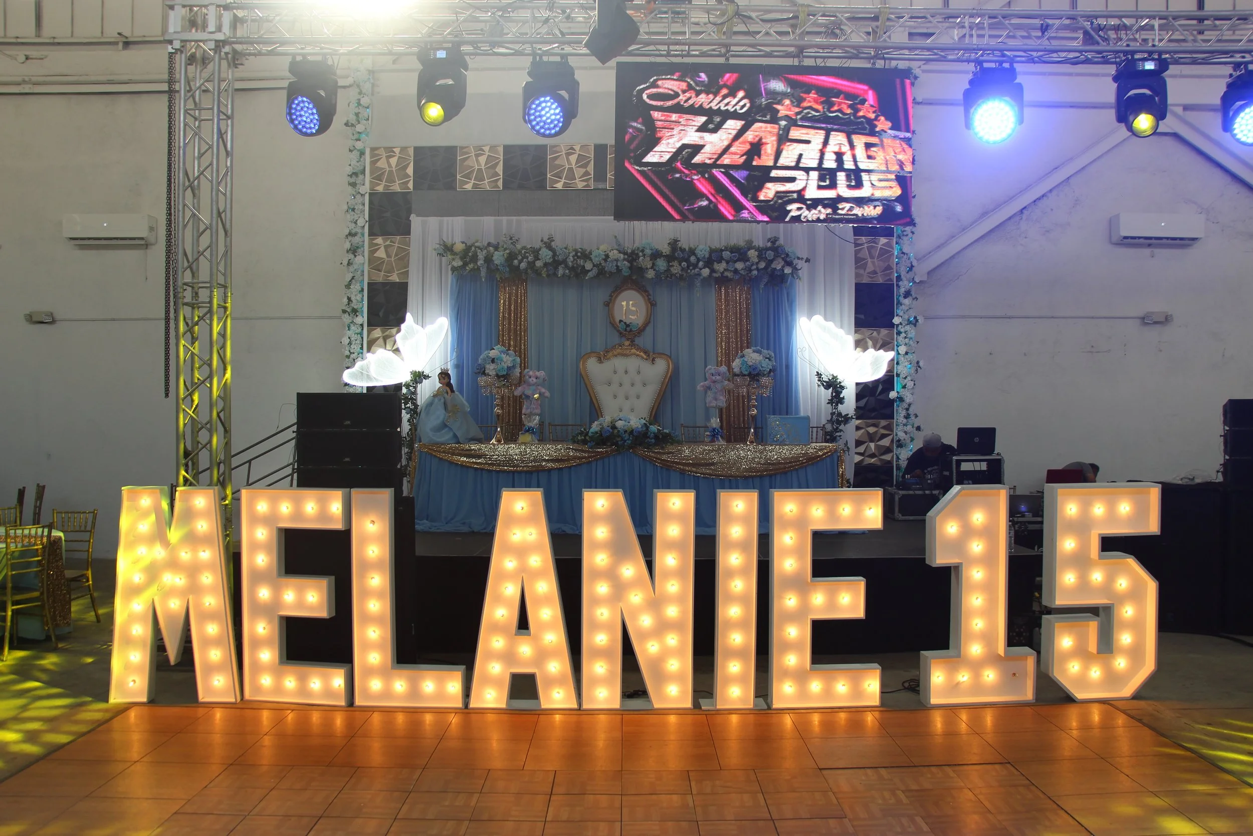 Decorated event stage with large illuminated letters spelling 'MELANIE 15', a chair in the center, blue and gold drapes, flower arrangements, and a digital screen displaying 'Sauda Takat Plus' at a 15th birthday celebration.