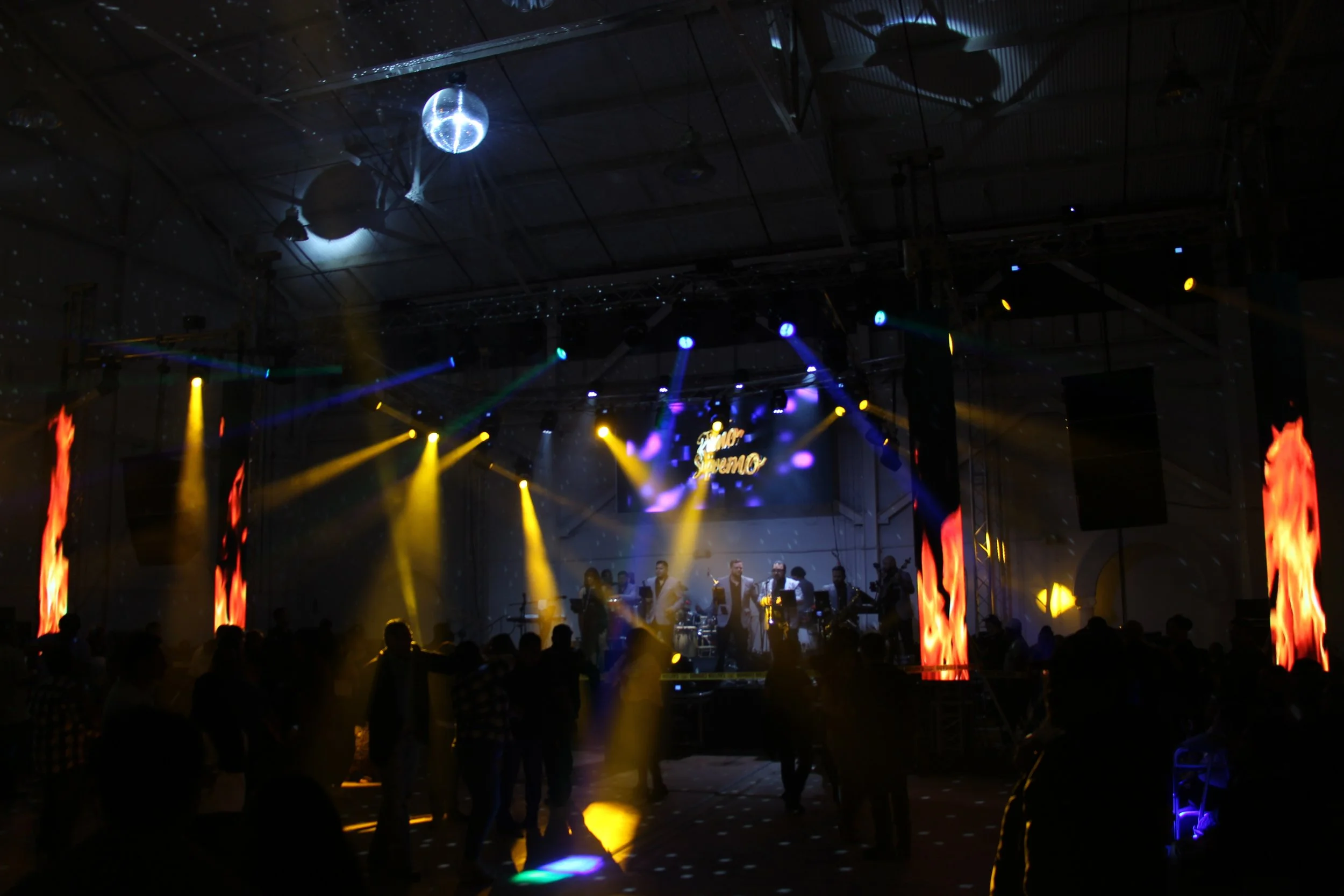 Indoor concert with a band on stage, bright colorful lights, disco ball reflections, and dancing crowd.