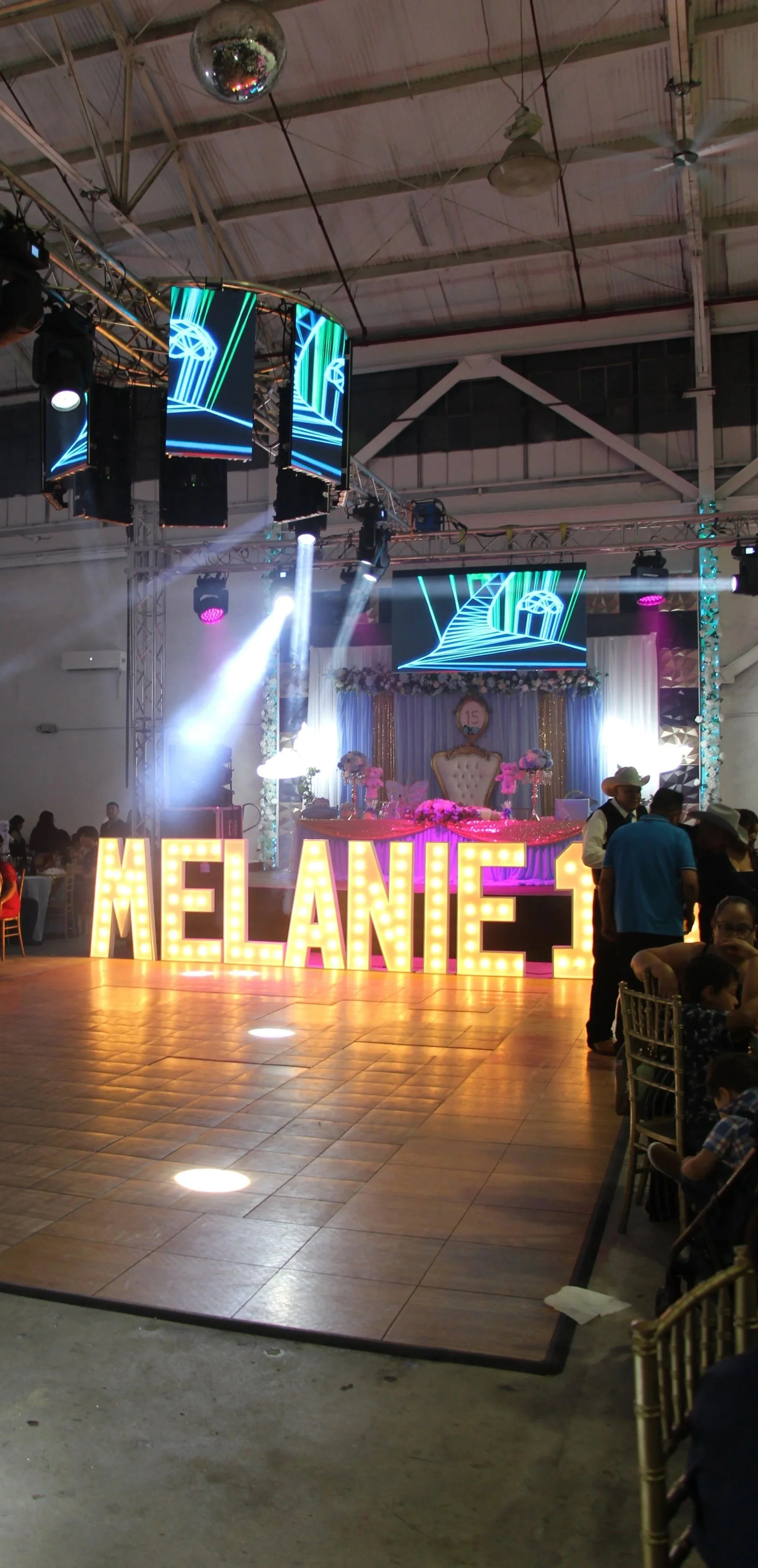 Decorated event stage with large illuminated letters spelling 'MELANIE 3', floral arrangements, a throne-like chair, and digital screens displaying neon graphics, with people gathered around in a festive indoor setting.
