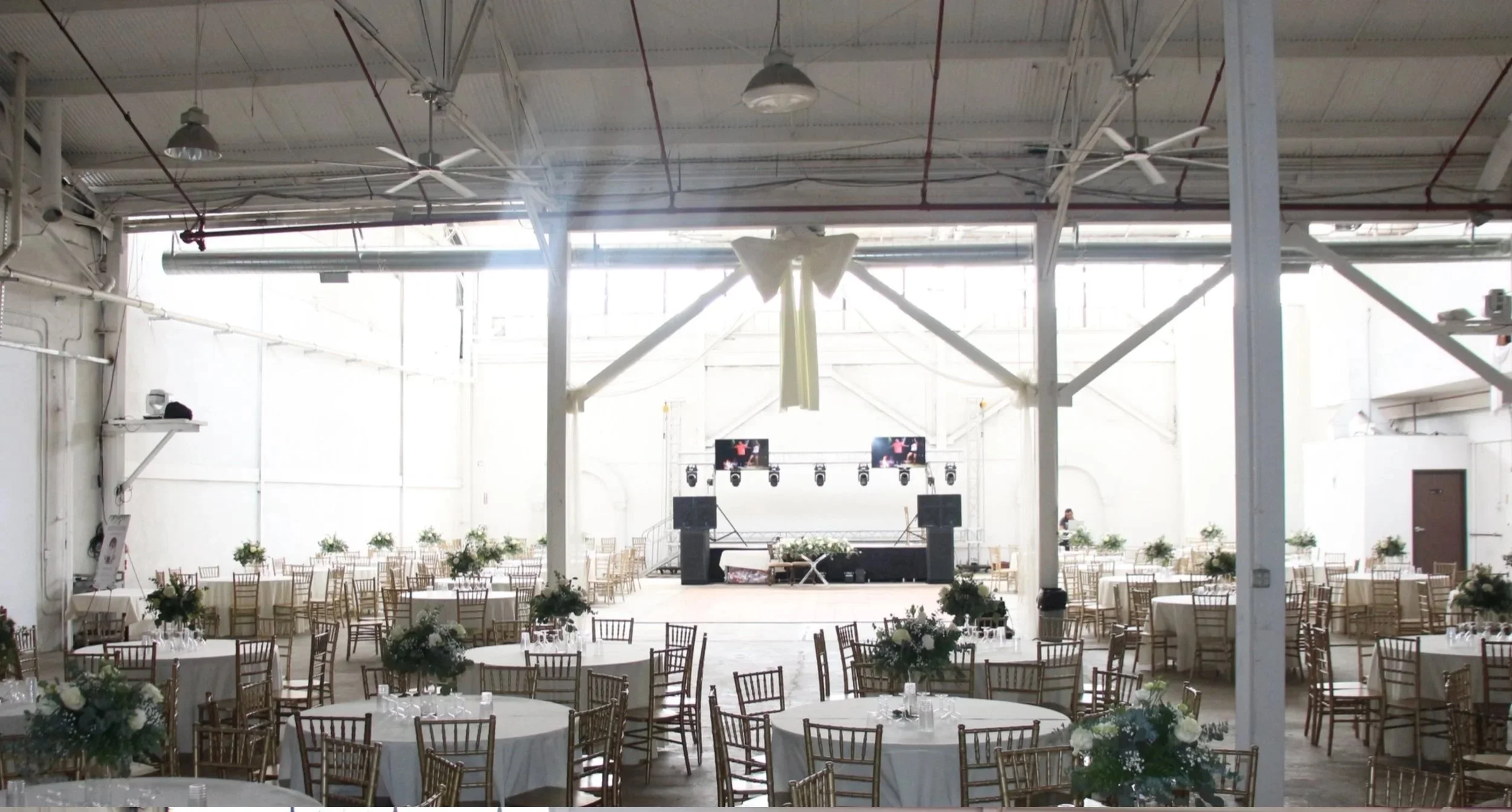 Empty event space with round tables decorated with floral centerpieces and gold chairs, a stage with lighting and screens, illuminated by natural light from large windows.