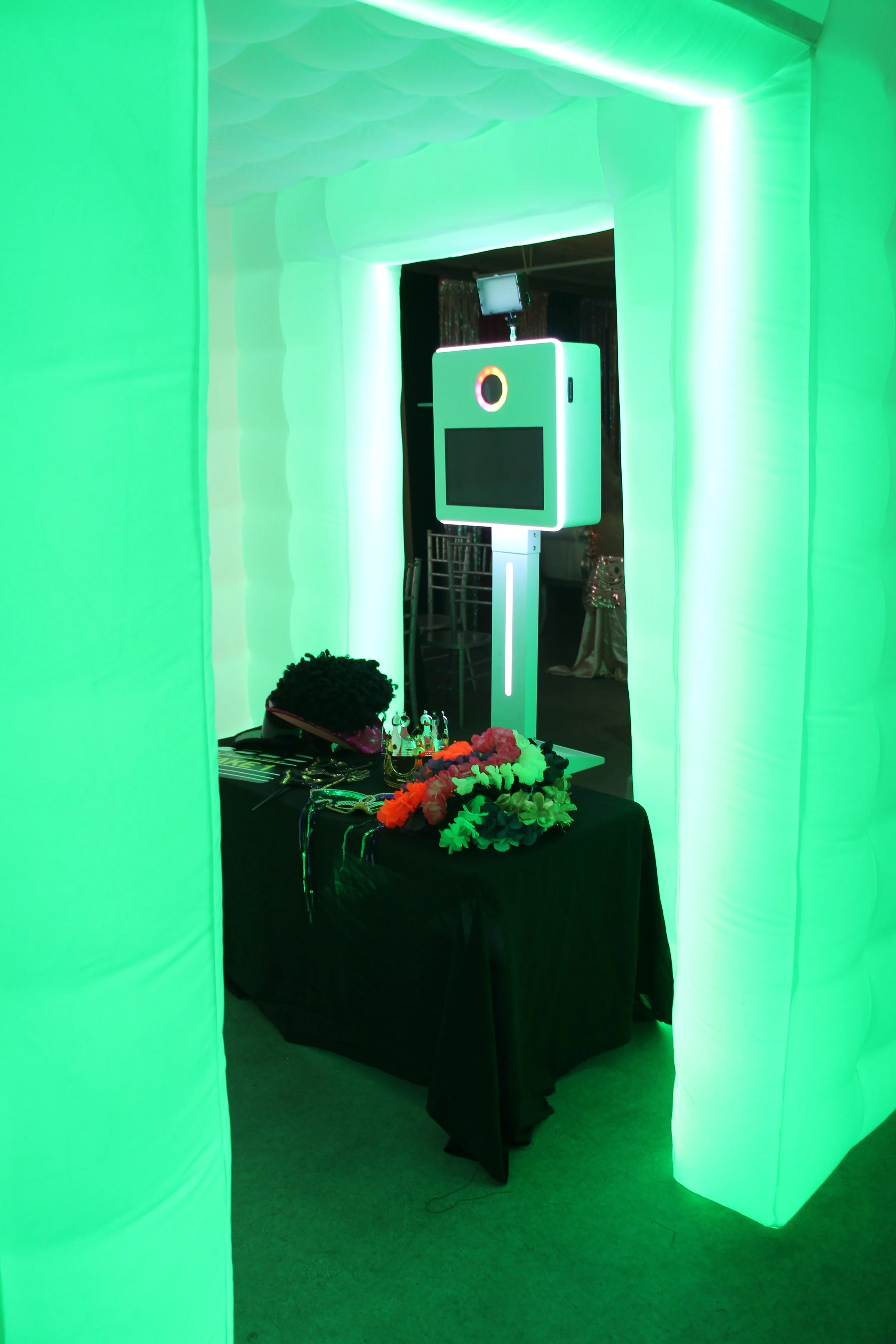 A photo booth with a green glowing inflatable structure, a table with leis and floral decorations, and a photo and video camera behind a screen.