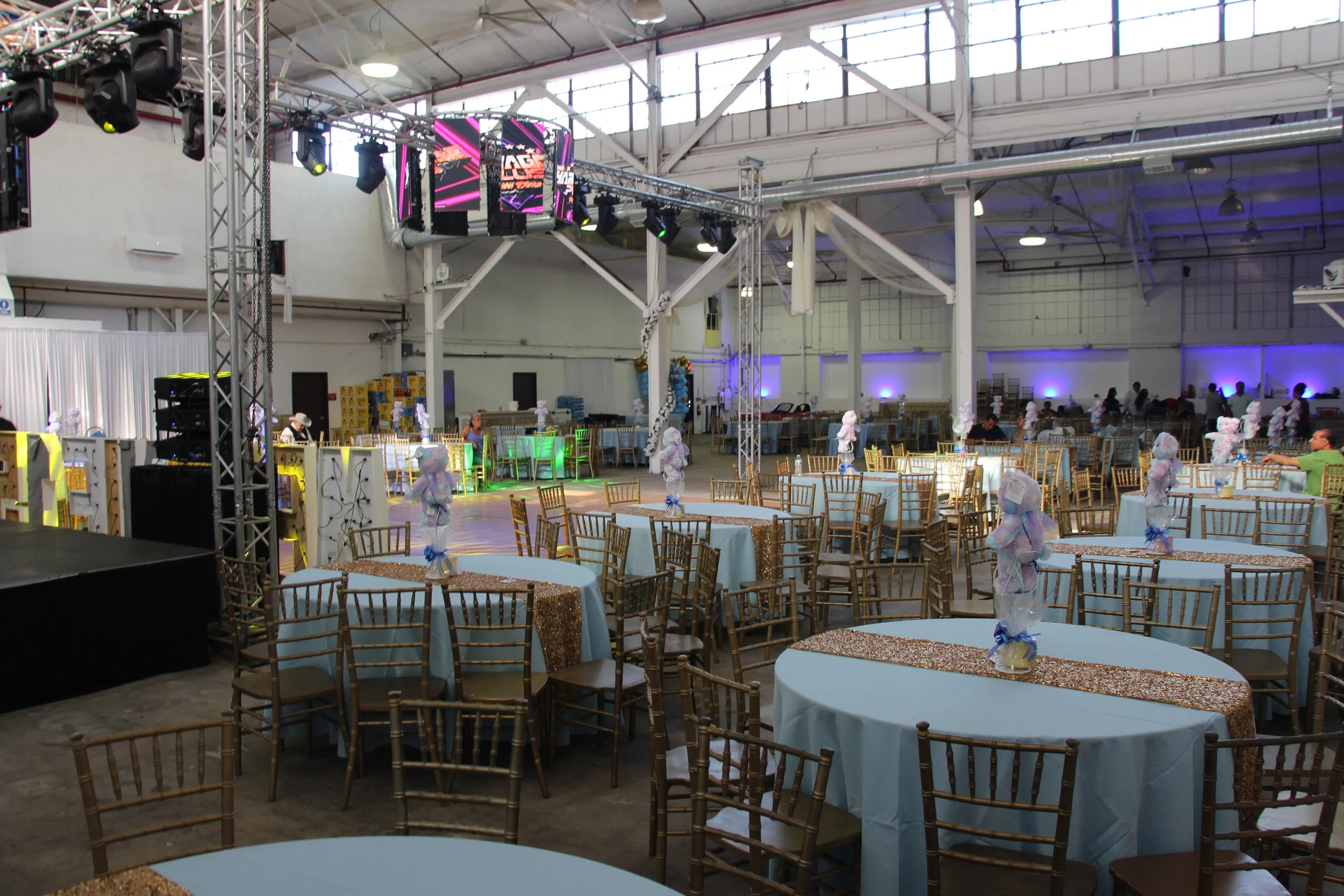 Event venue with round tables covered in white tablecloths and decorated with small centerpiece figures, gold Chiavari chairs, a stage with lighting and a large screen, and a spacious industrial-style ceiling with rigging.