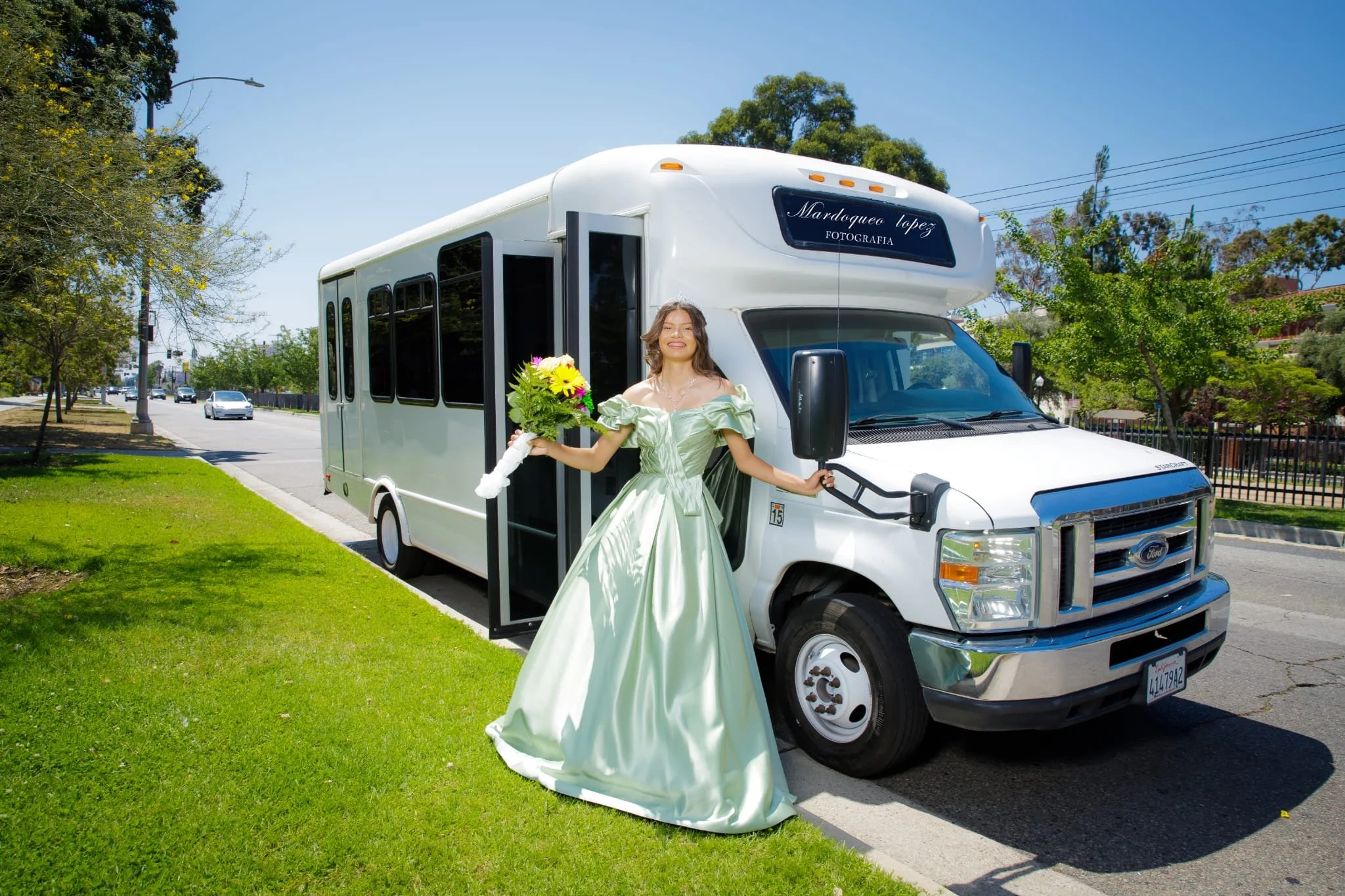 A woman in a green dress holding a bouquet of flowers standing in front of a white shuttle bus on a sunny street with trees and cars in the background.