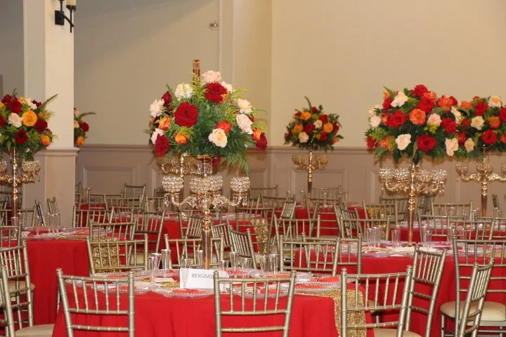Elegant banquet hall decorated with tall flower arrangements on crystal stands and red tablecloths covering round tables with place settings, and gold chairs arranged around the tables.