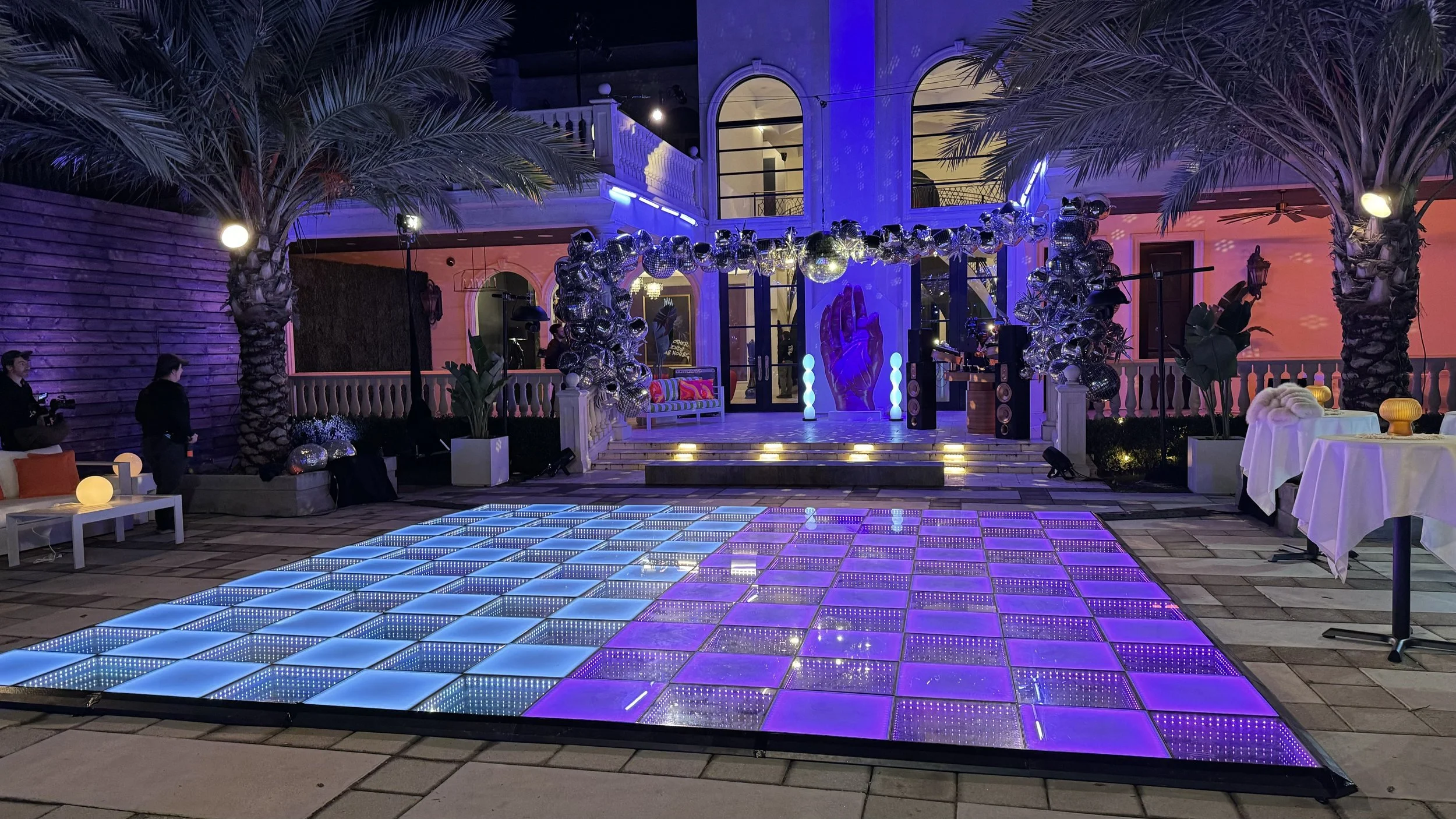 Outdoor party scene with a illuminated dance floor in blue and purple, palm trees, a stage with a balloon decoration arch, and seating areas with tables and couches at night.