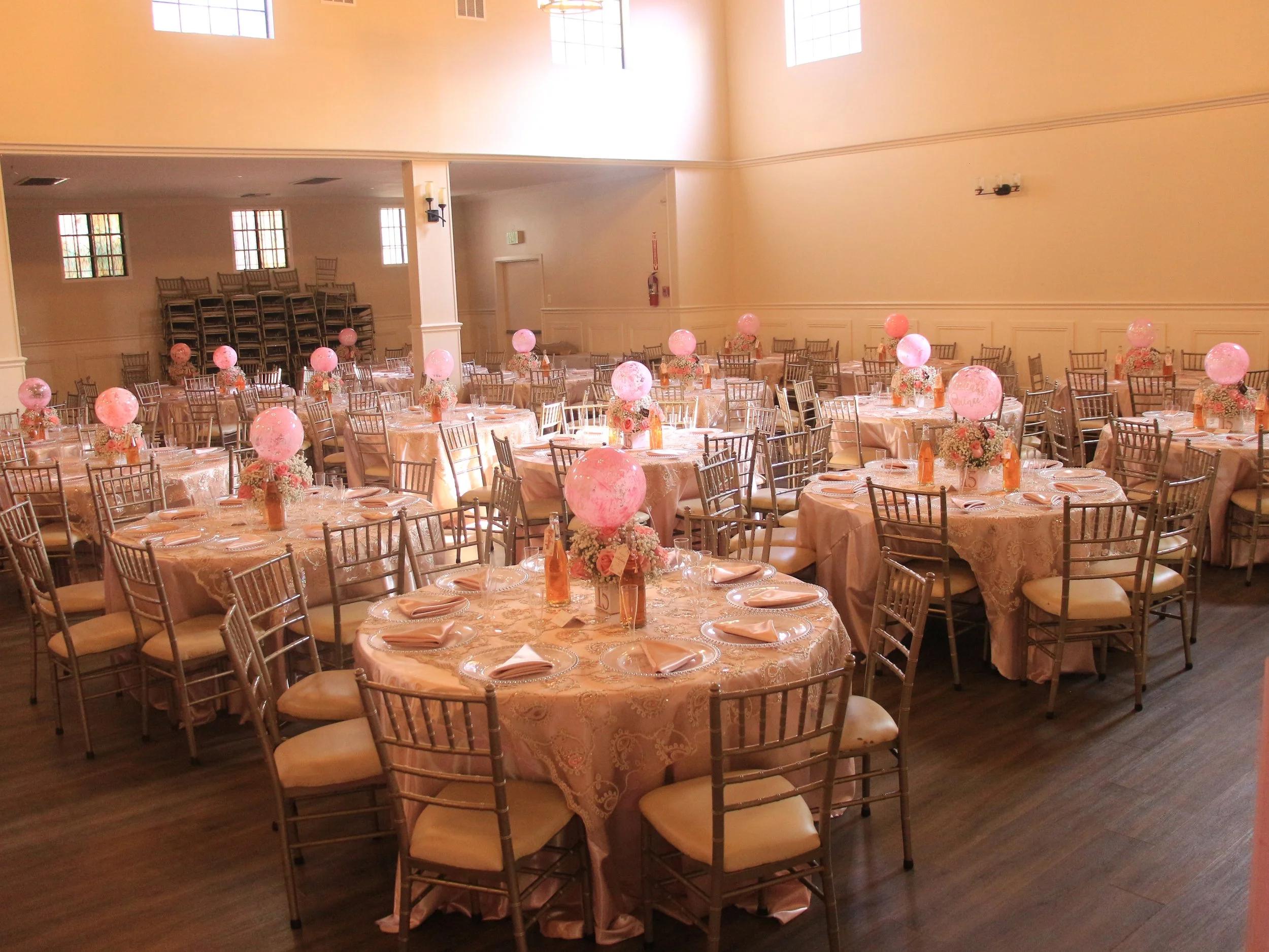 A banquet hall decorated with round tables covered with pink tablecloths, each with a bouquet of flowers and pink balloons as centerpieces, arranged for a celebration or wedding reception.