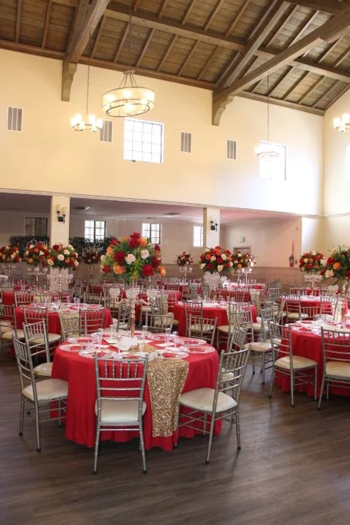 Wedding reception with round tables draped in red tablecloths, decorated with large floral centerpieces of red and pink flowers, and silver chairs, in a spacious hall with high ceiling and wooden beams.