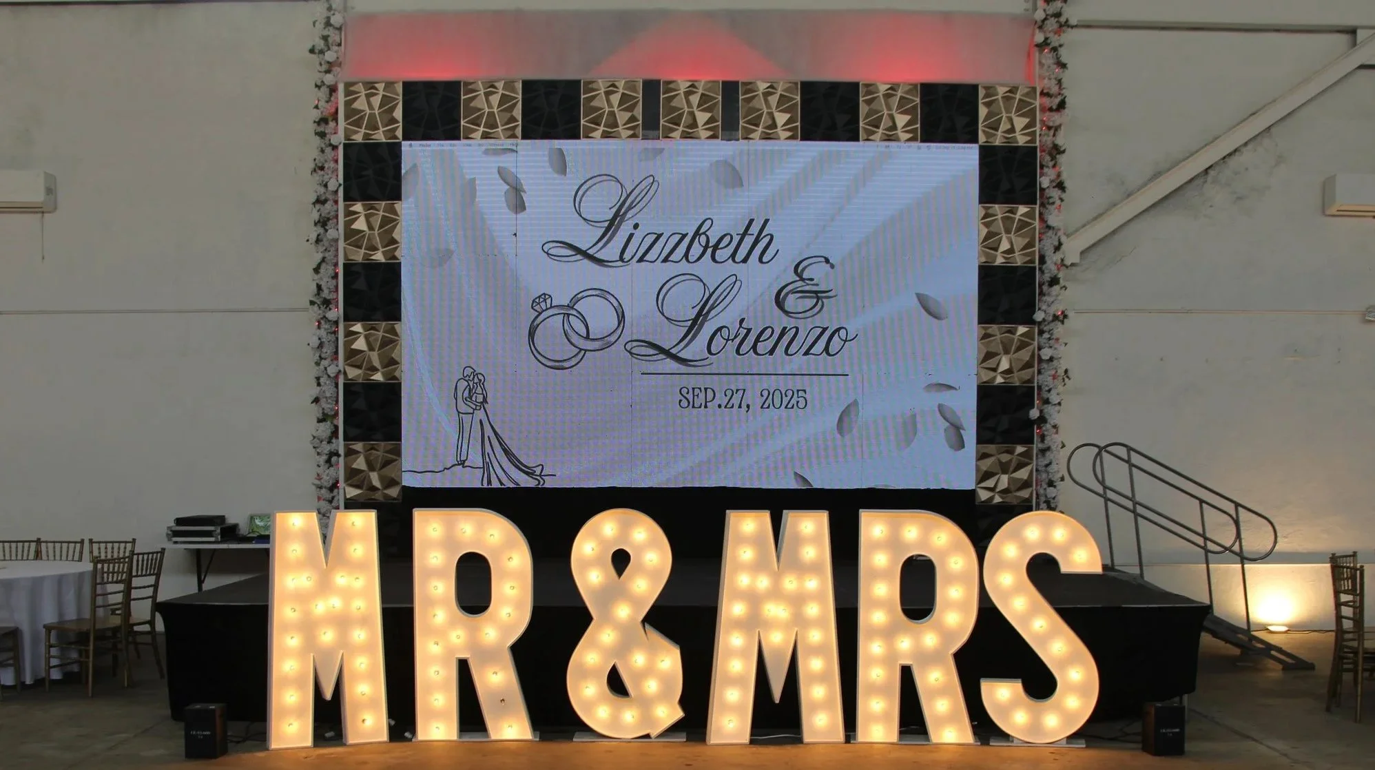 Wedding reception setup with illuminated large letters spelling 'MR & MRS' in front of a large digital display featuring the names Lizbeth and Lorenzo, wedding date September 27, 2025, and wedding illustrations, decorated with flowers and soft lighting.