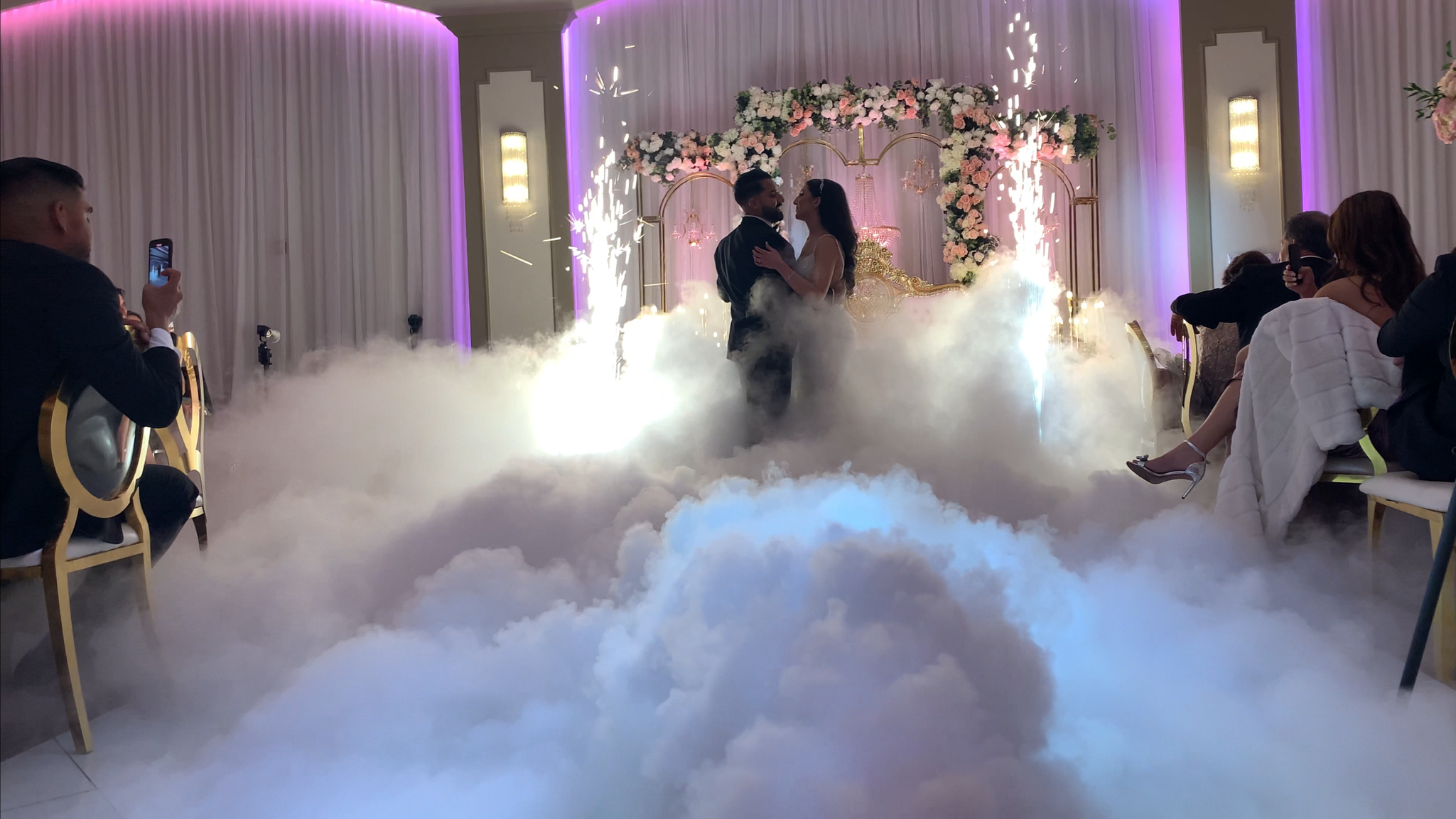 A bride and groom are dancing in the center of a wedding reception, surrounded by artificial fog and fireworks, with seated guests watching and taking photos.