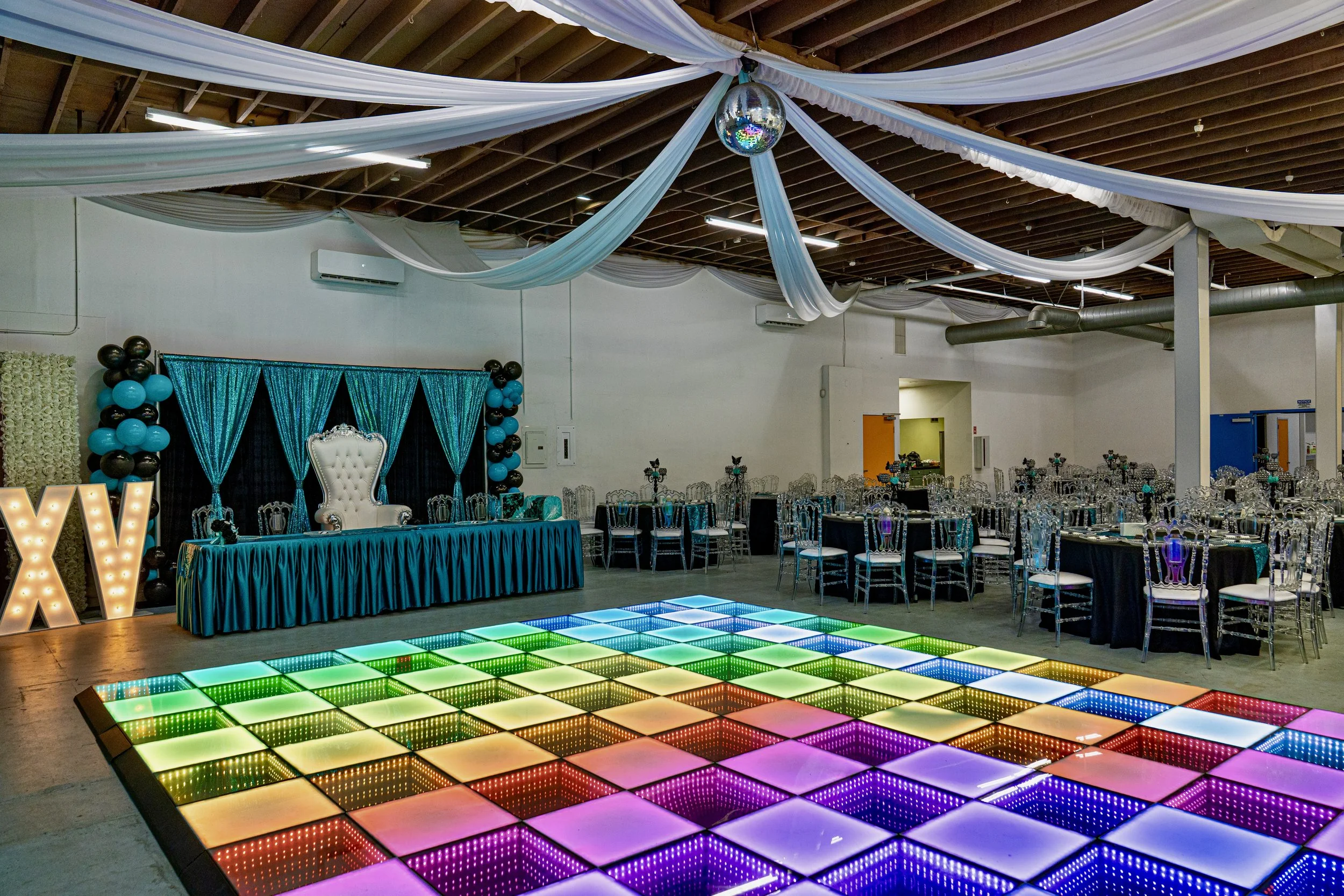 Decorated event space with draped fabric on the ceiling, illuminated dance floor with colorful lights, a decorated backdrop with blue curtains and balloons, a white throne-like chair on a table, and tables with silver chairs for guests.