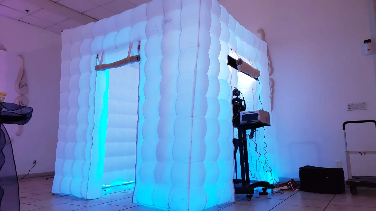 An inflatable photo booth with a glowing interior, set up indoors, with equipment such as a projector and lights around it.
