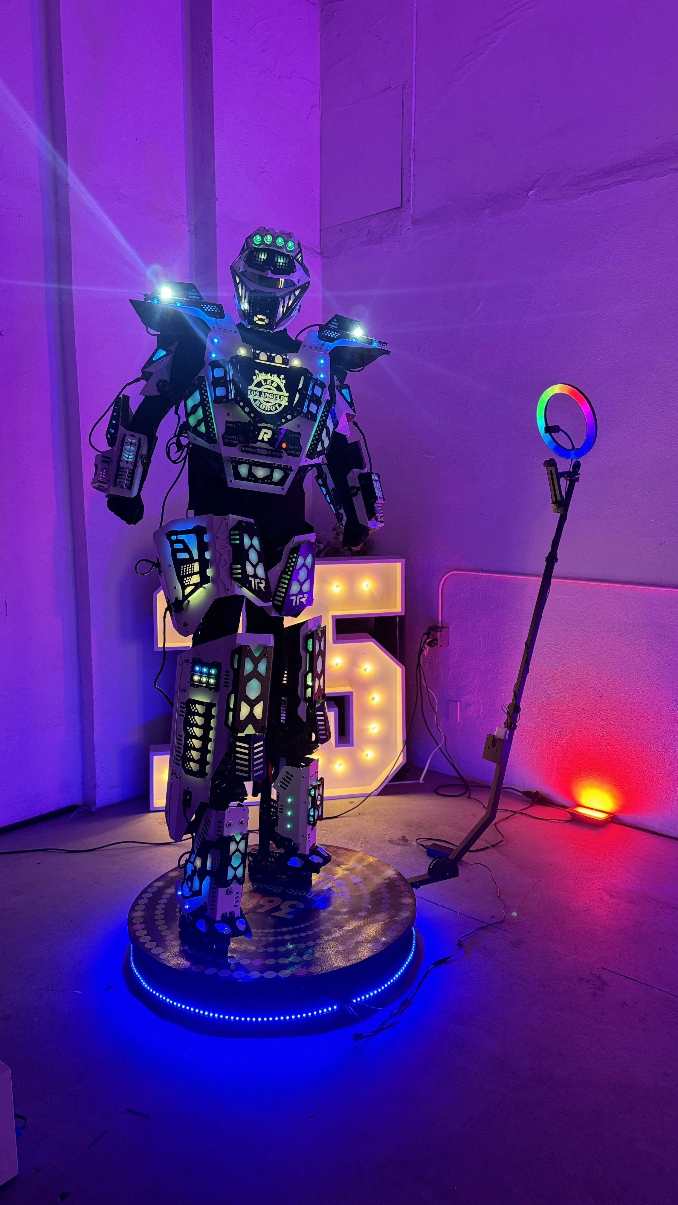A humanoid robot with LED lights standing on a circular platform in a room with colorful neon lighting, a large illuminated number 5 in the background, and a robot-themed ring light on a stand.