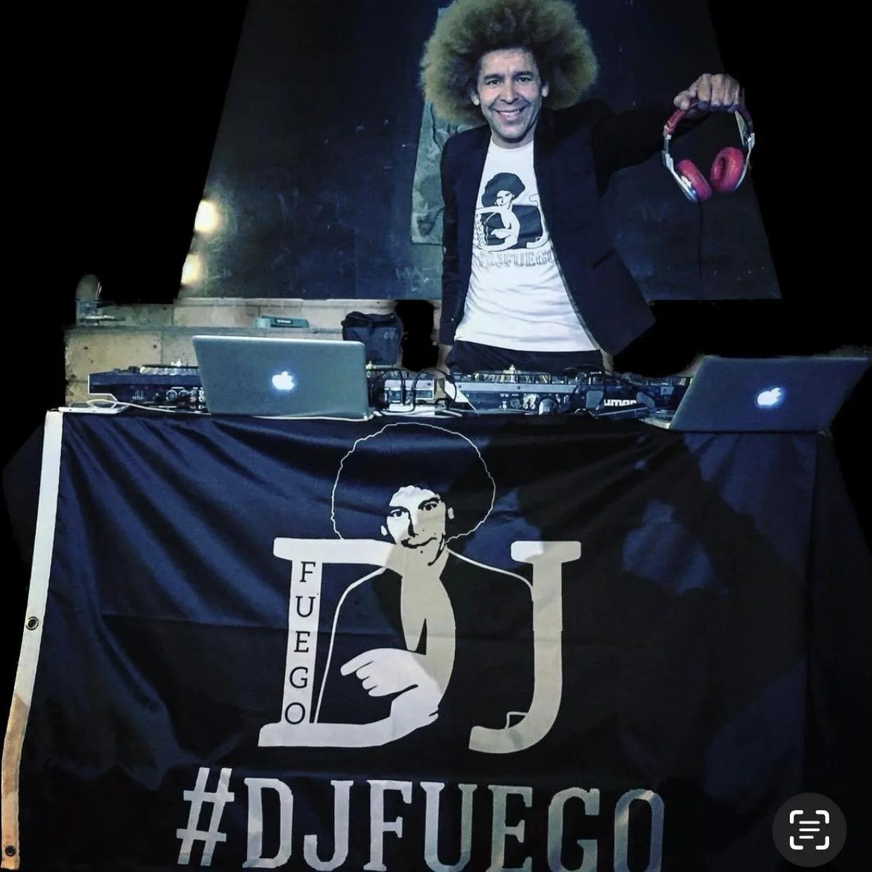 A DJ with an afro hairstyle standing behind a DJ booth, smiling and holding pink headphones in one hand. The booth has a banner with a drawing of a person with an afro and the words "FUEGO DJ" and "#DJFUEGO." The DJ is wearing a graphic T-shirt and a dark jacket, with laptop computers on the booth.