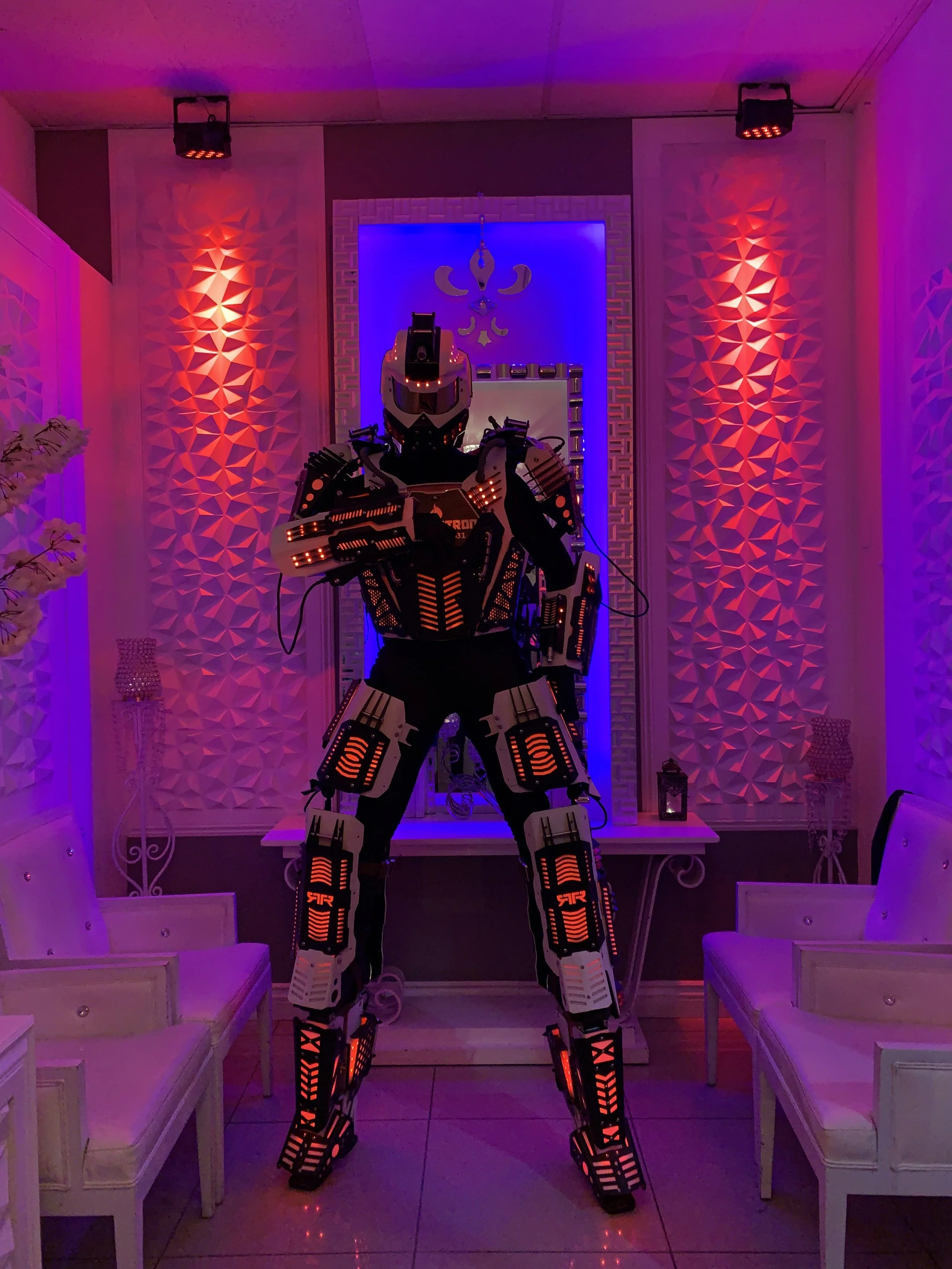 A person in a futuristic, black and white robotic suit with orange LED lights stands inside a room with purple and pink lighting, white chairs, decorative wall panels, and a small table with lanterns.