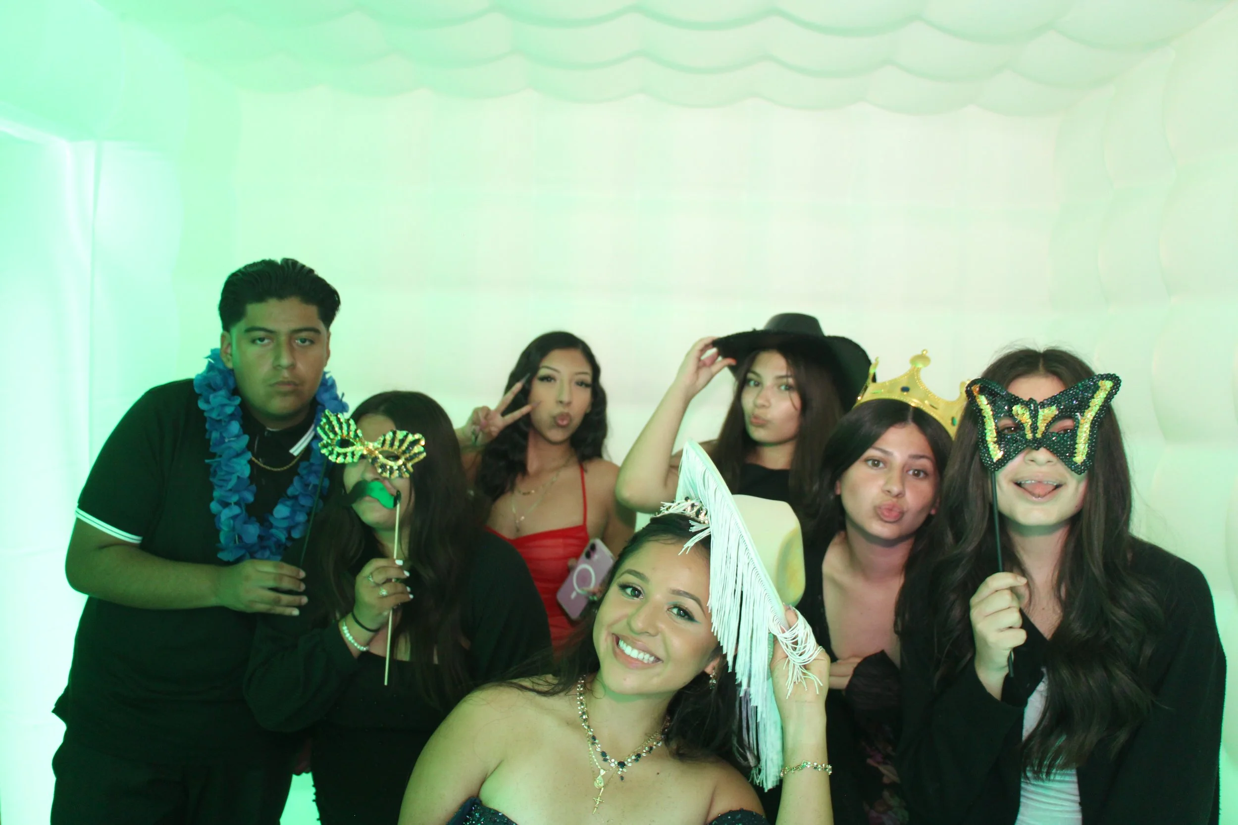 Group of seven people at a party, wearing costumes and accessories, posing for a photo with some holding masks, tiaras, and wearing hats, in front of a green-lit background.