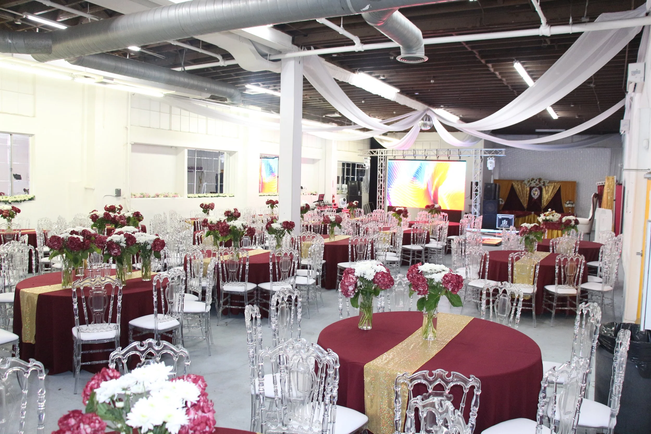 Indoor event space decorated with burgundy tablecloths, gold runners, and floral centerpieces of pink and white flowers in glass vases. Transparent chairs surround the tables. There's a stage with large screens and lighting equipment at the front of the room. White draped fabric decorates the ceiling.