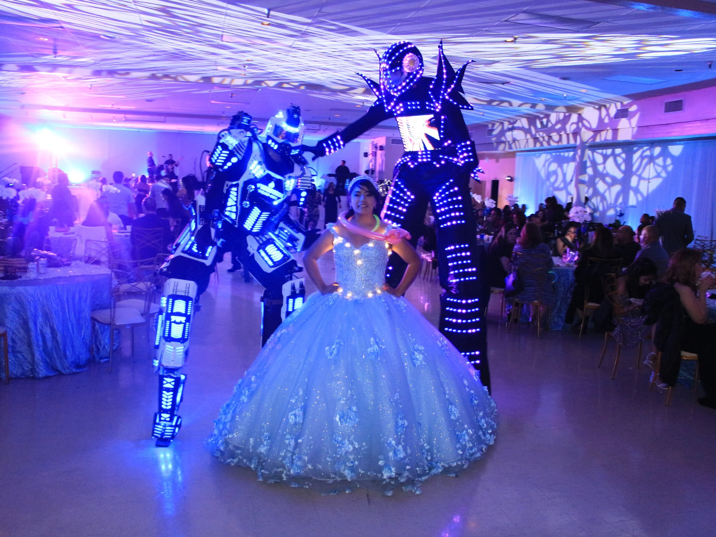A woman in a white, glittery ball gown stands between two individuals dressed in illuminated robot costumes at a lively event or party with many guests seated at tables and colorful lighting.