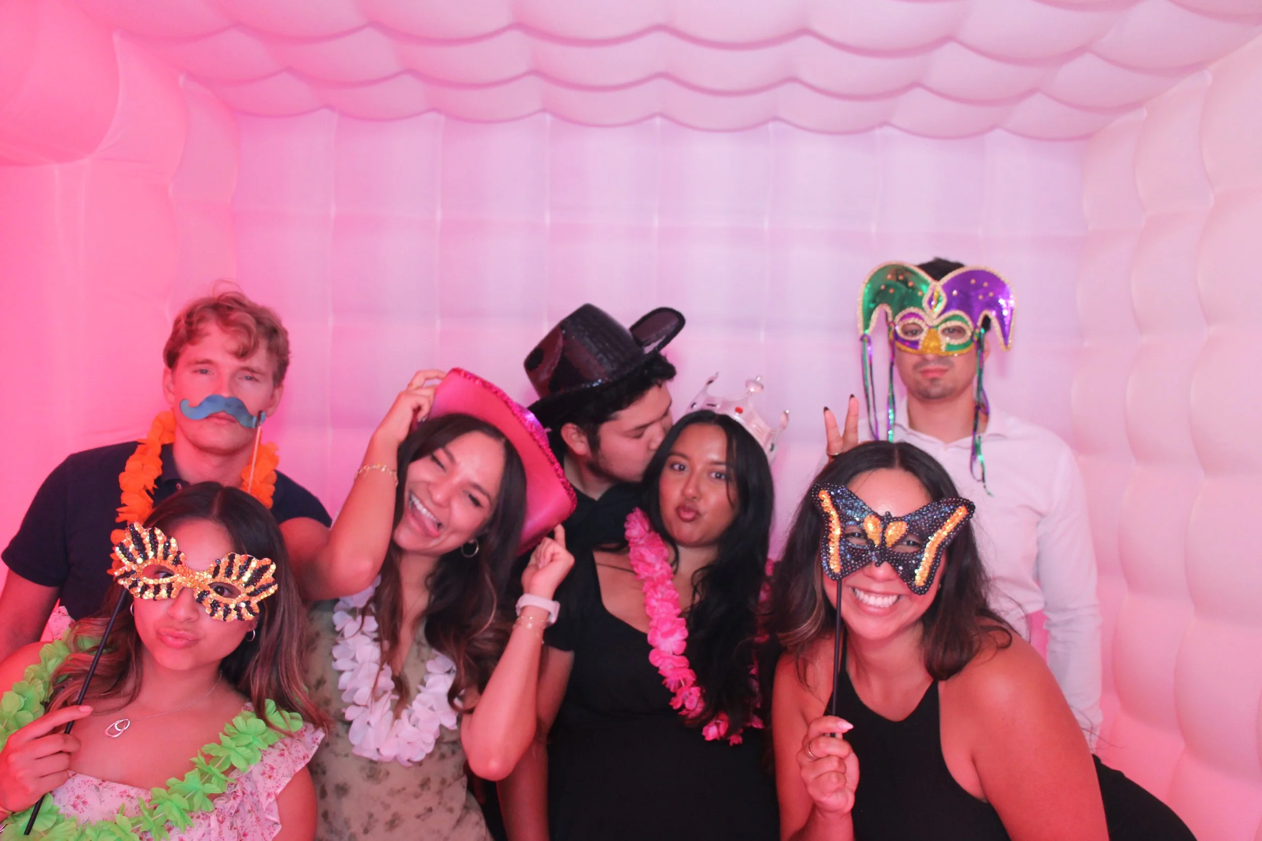 Group of people wearing colorful masks and hats at a party in a pink inflatable room.