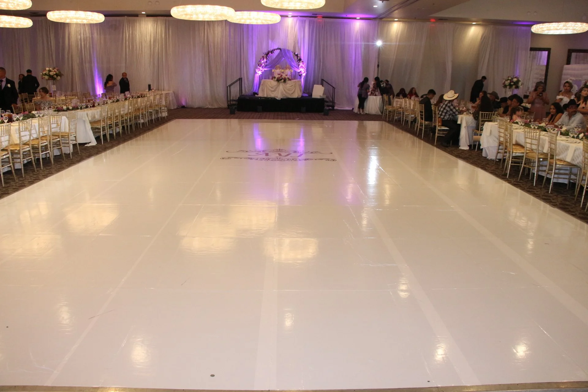 Large decorated event hall with a dance floor in the center, surrounded by banquet tables with chairs, and an elevated stage with a floral arch at the far end. People are sitting and standing around the tables.