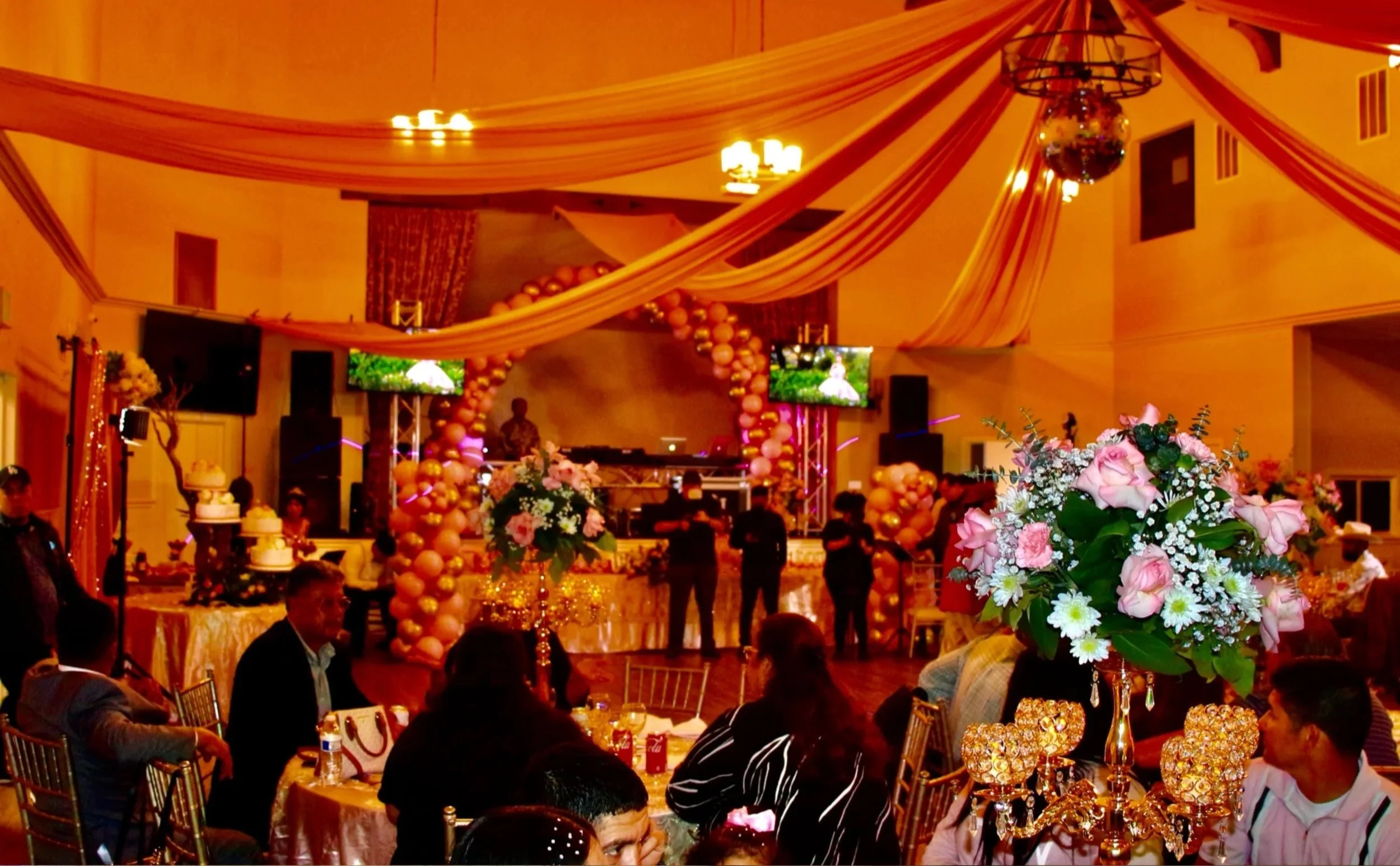 Decorated banquet hall with draped fabric, balloon arch, floral centerpieces, and a stage with questions.