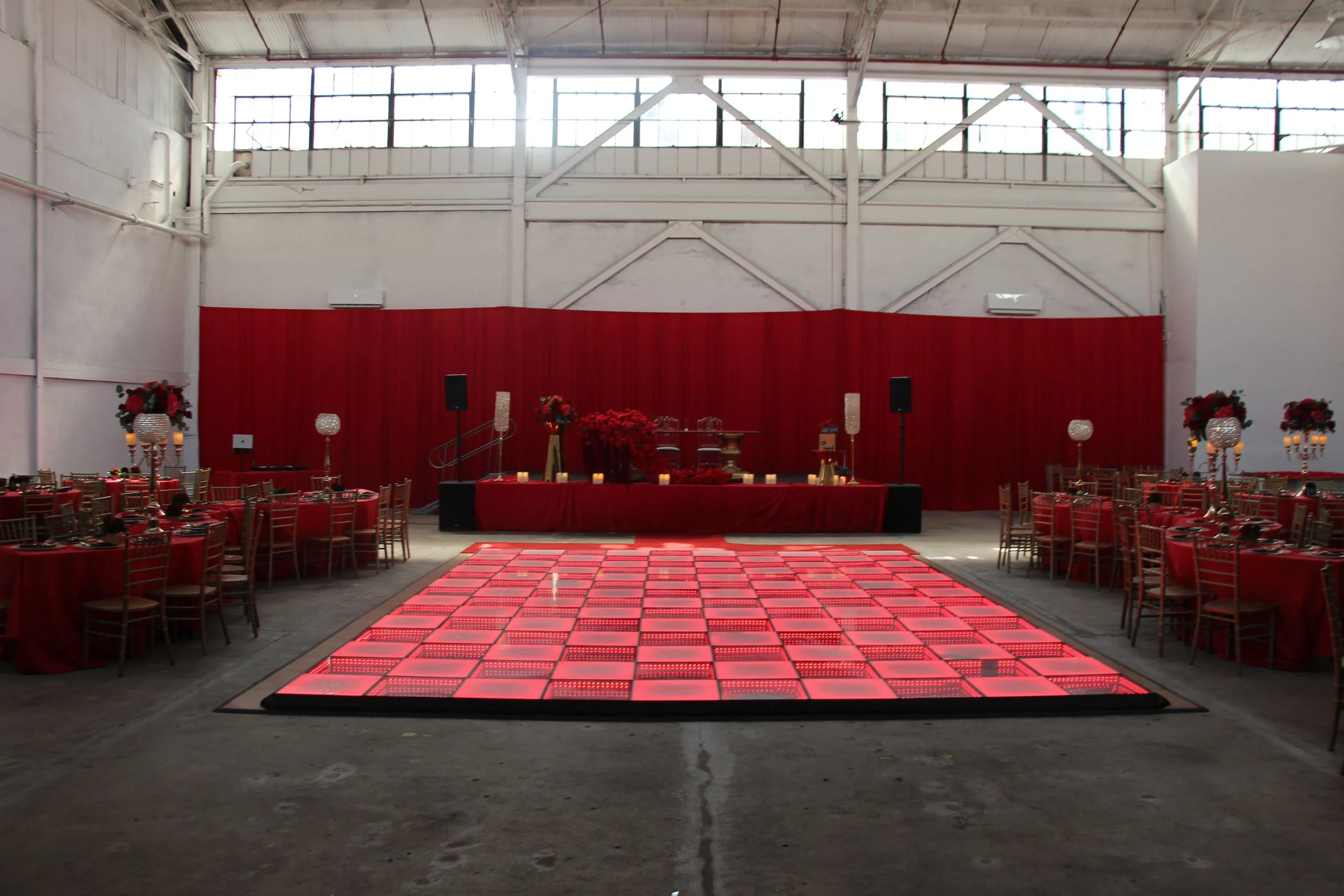 Indoor event space decorated in red with tables, chairs, floral arrangements, and a lit dance floor.