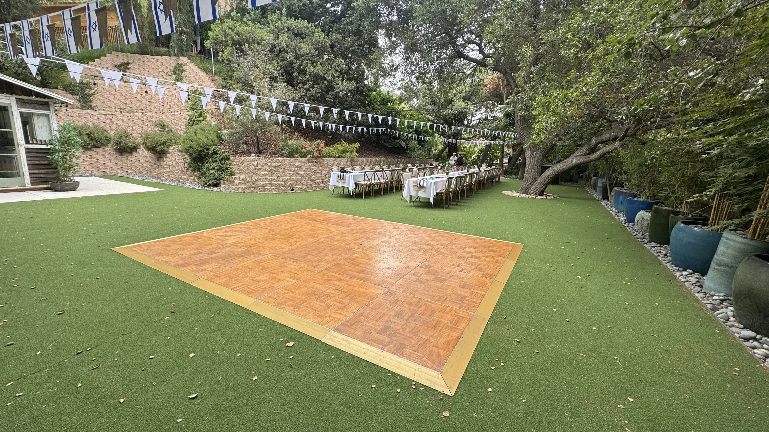 Outdoor backyard with green artificial grass, a small wooden dance floor, and a long dining table set with white tablecloths, surrounded by potted plants and large trees. Blue and white flags are strung overhead.