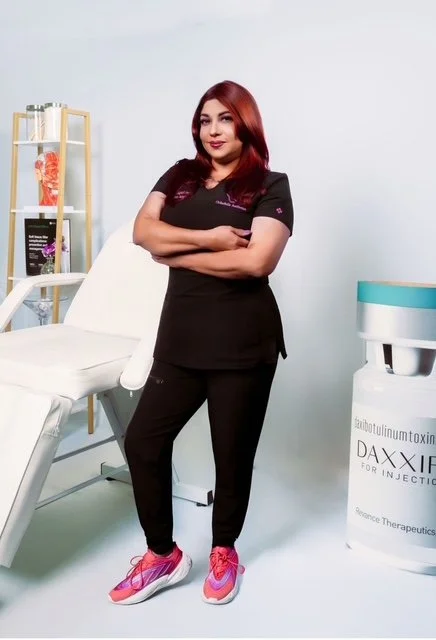 A woman standing in a medical or skincare clinic with her arms crossed, wearing black scrubs and pink sneakers. There is a white examination chair and medical equipment in the background.