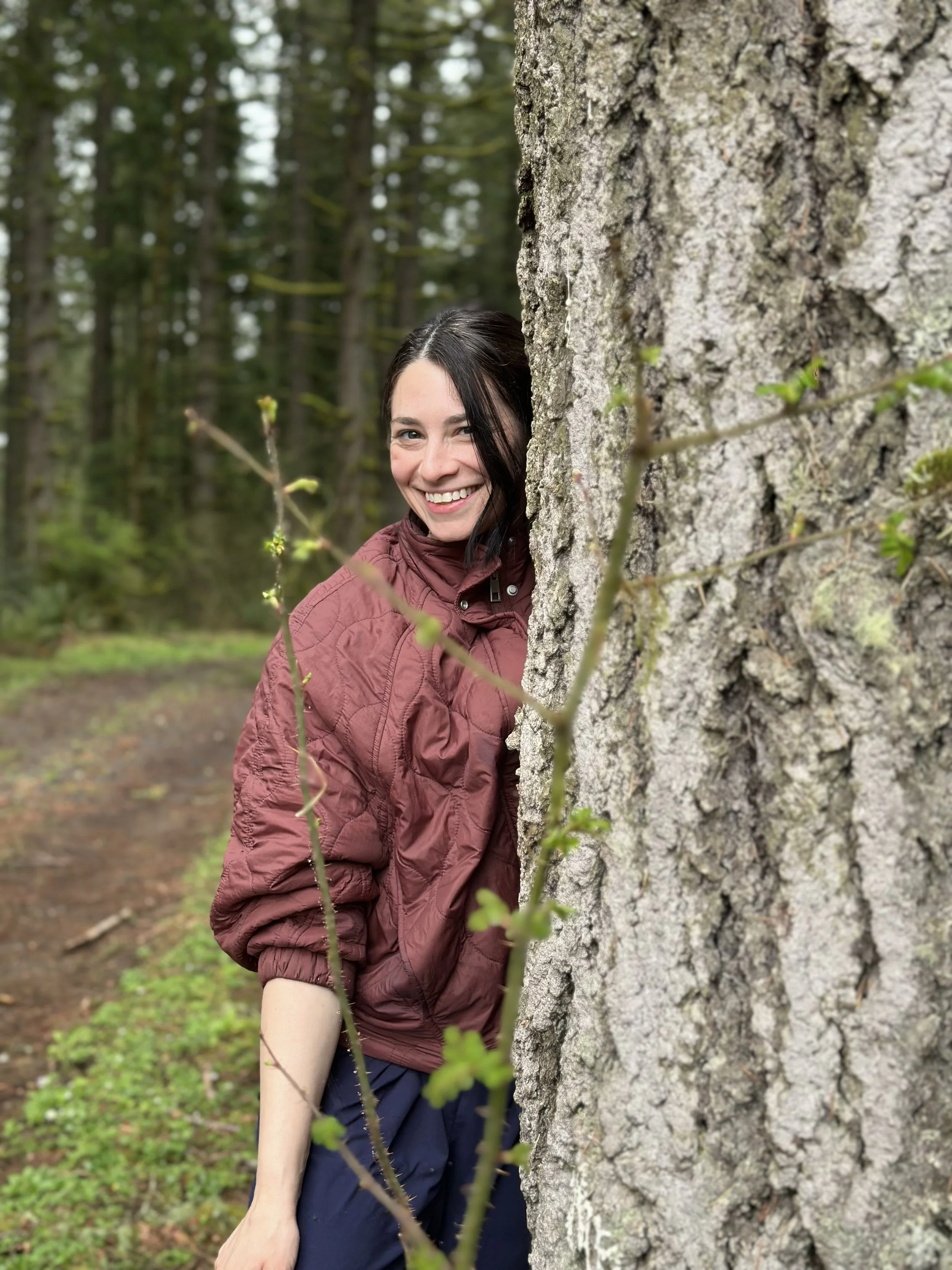 A woman with dark hair smiling and peeking from behind a large tree trunk in a forest. She is wearing a maroon jacket and blue pants, with green foliage and tall trees in the background.