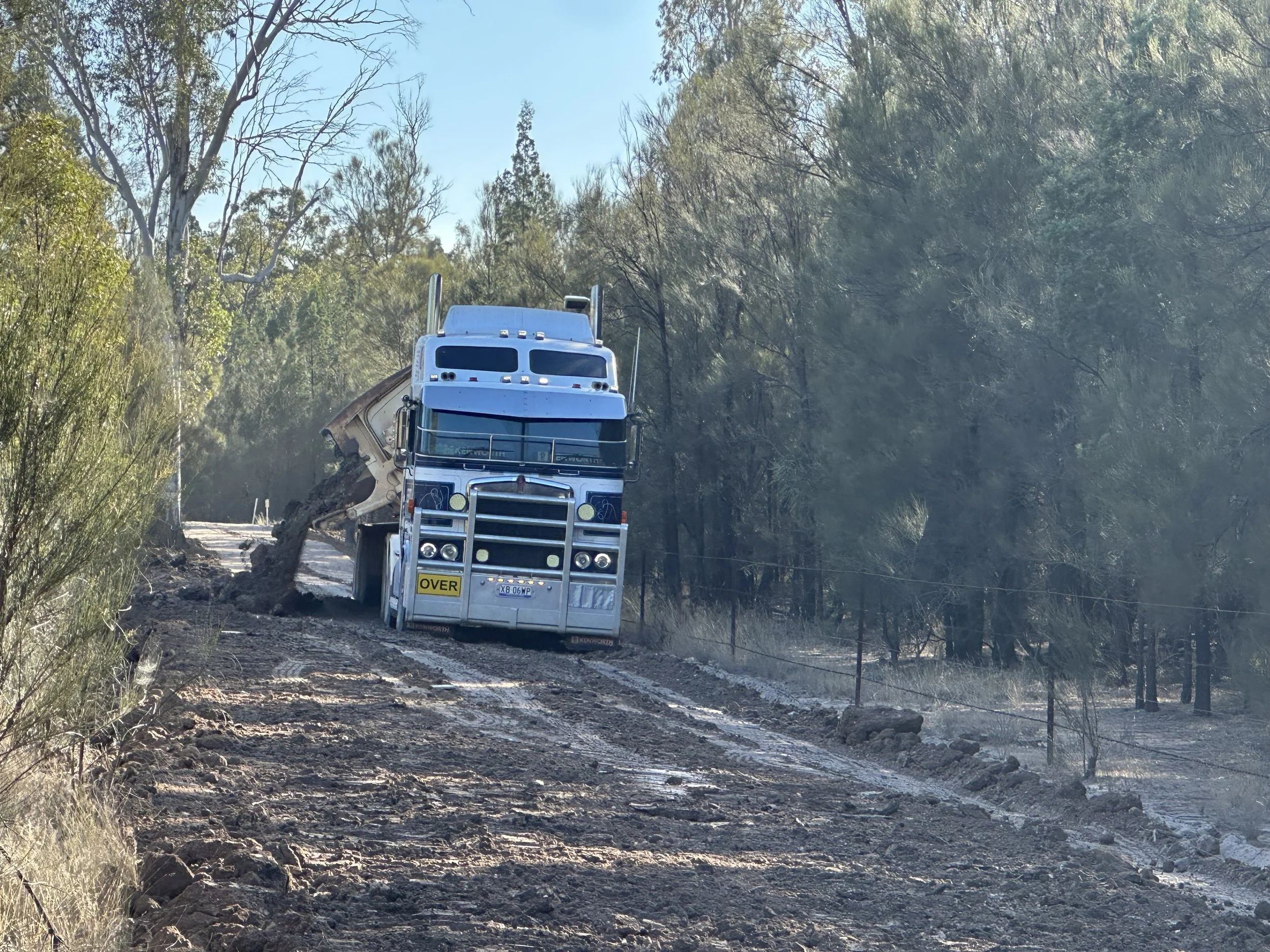 A large semi-truck driving on a dirt road through a forested area, with a construction or dirt clearing site showing disturbed soil and smoke in the background.