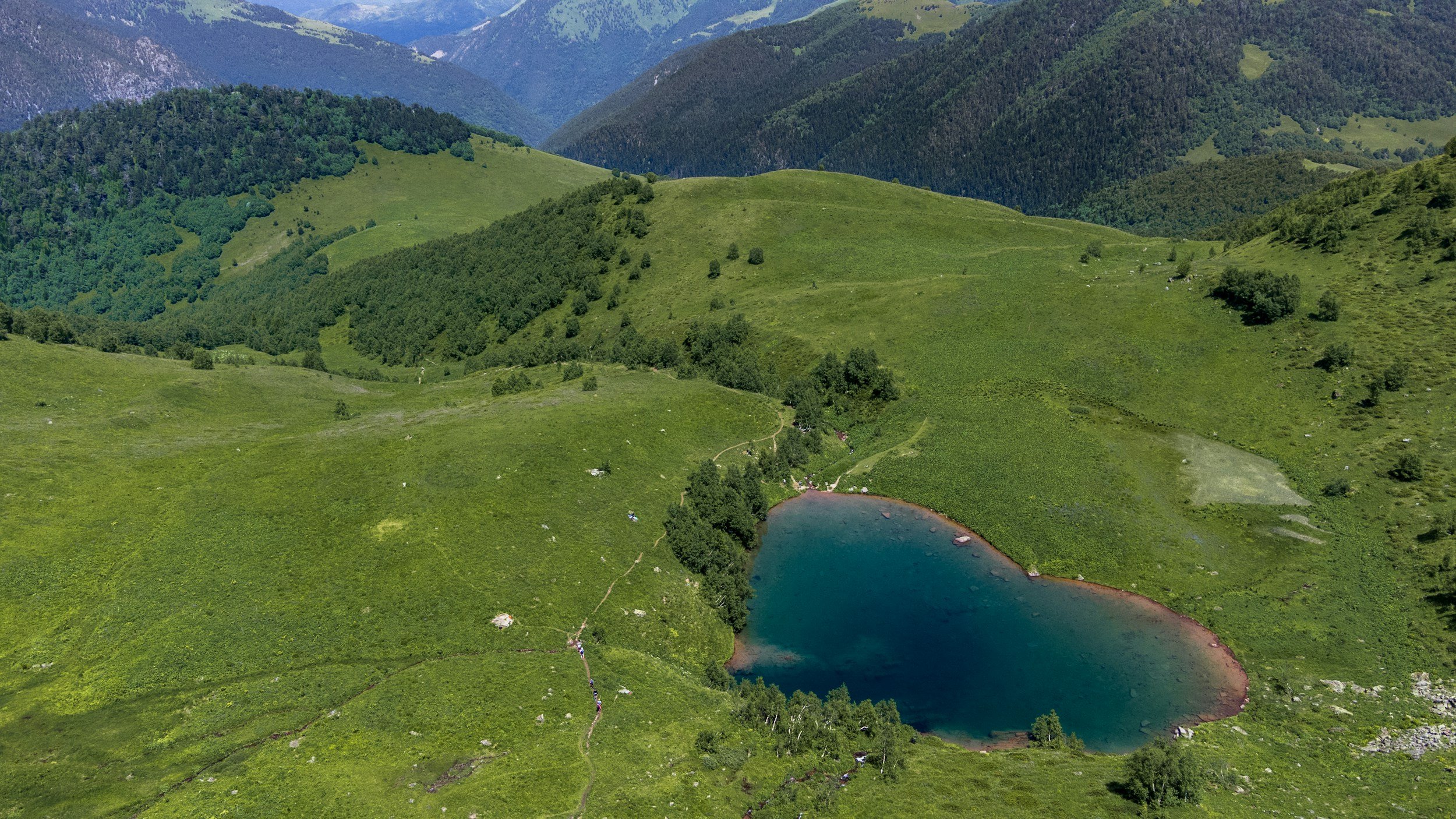 Aerial view of a green mountainous landscape with a small lake at the base, surrounded by grassy hills and patches of forest under a partly cloudy sky.