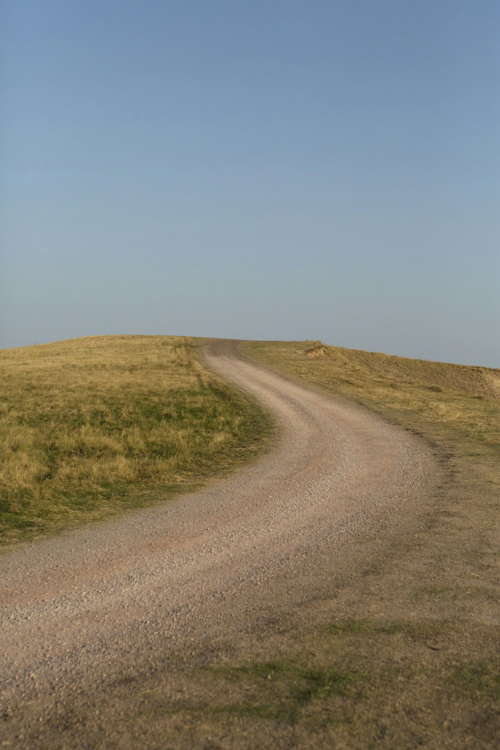 A gravel dirt road winding over grassy hills under a blue sky.