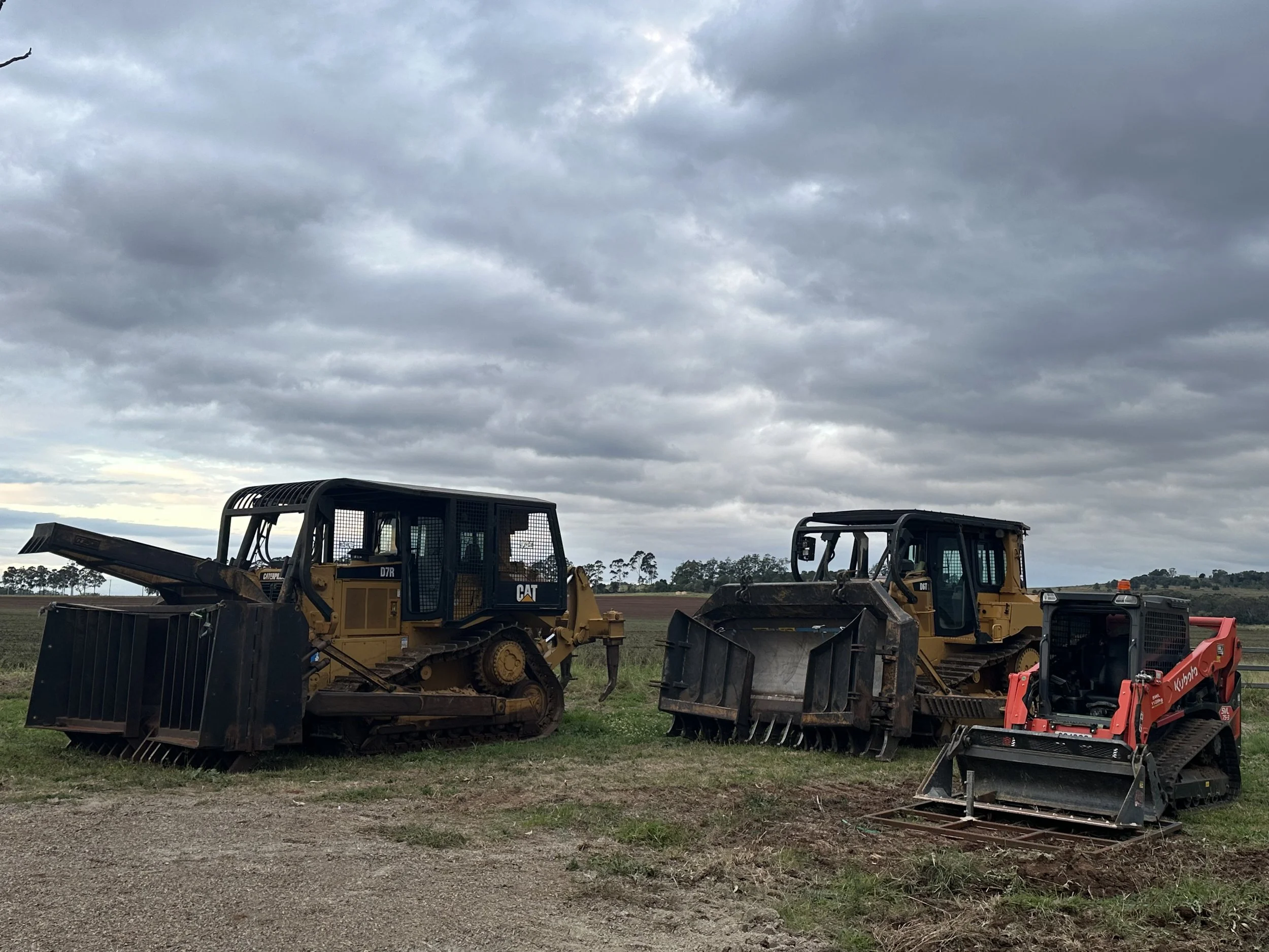 Three construction vehicles parked on grass under cloudy sky, two yellow bulldozers and a red mini loader.
