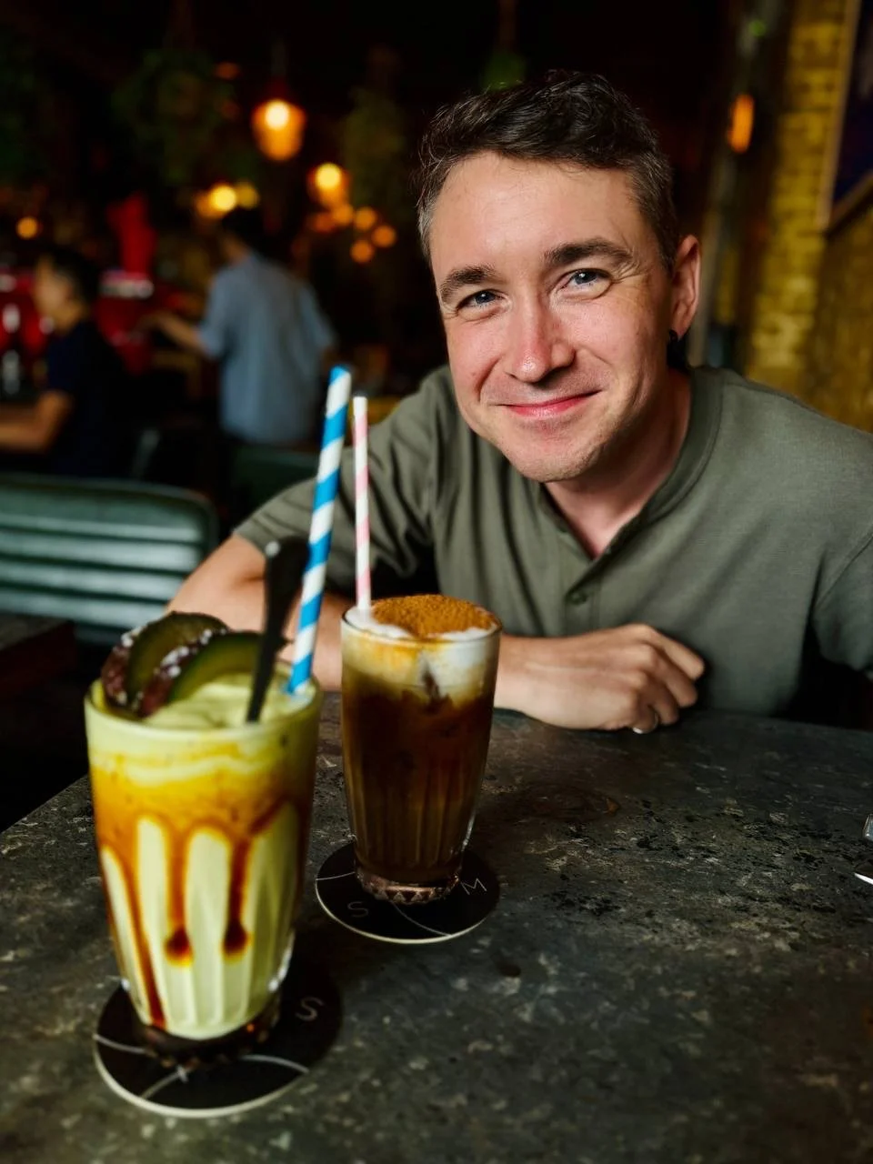 A smiling man sitting at a bar table with two colorful milkshakes, one topped with whipped cream and a cinnamon or nut sprinkle, in front of him.