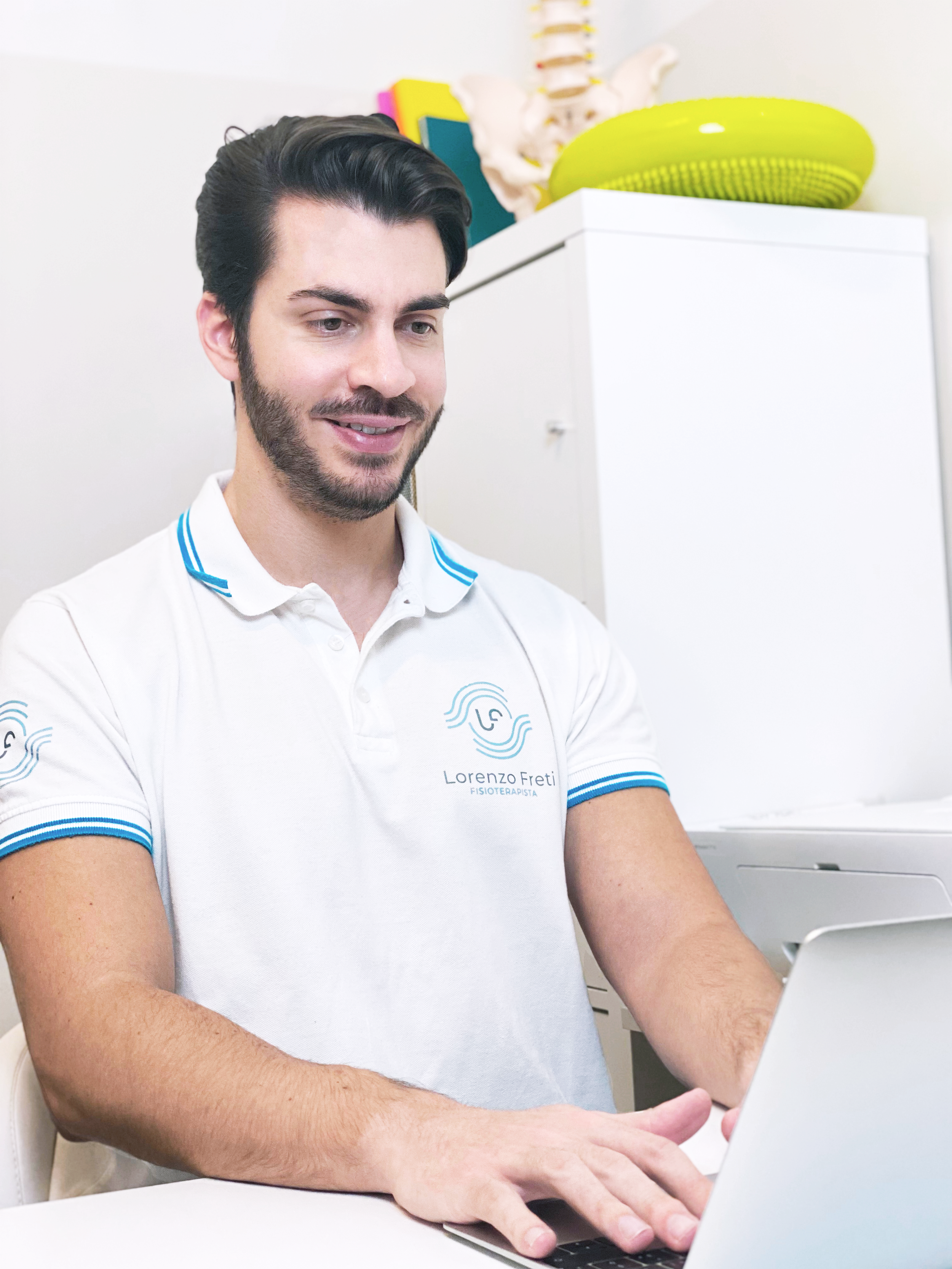 Un uomo con camicia bianca con scritta Lorenzo Freti, fisioterapista, che lavora su un laptop in uno studio.