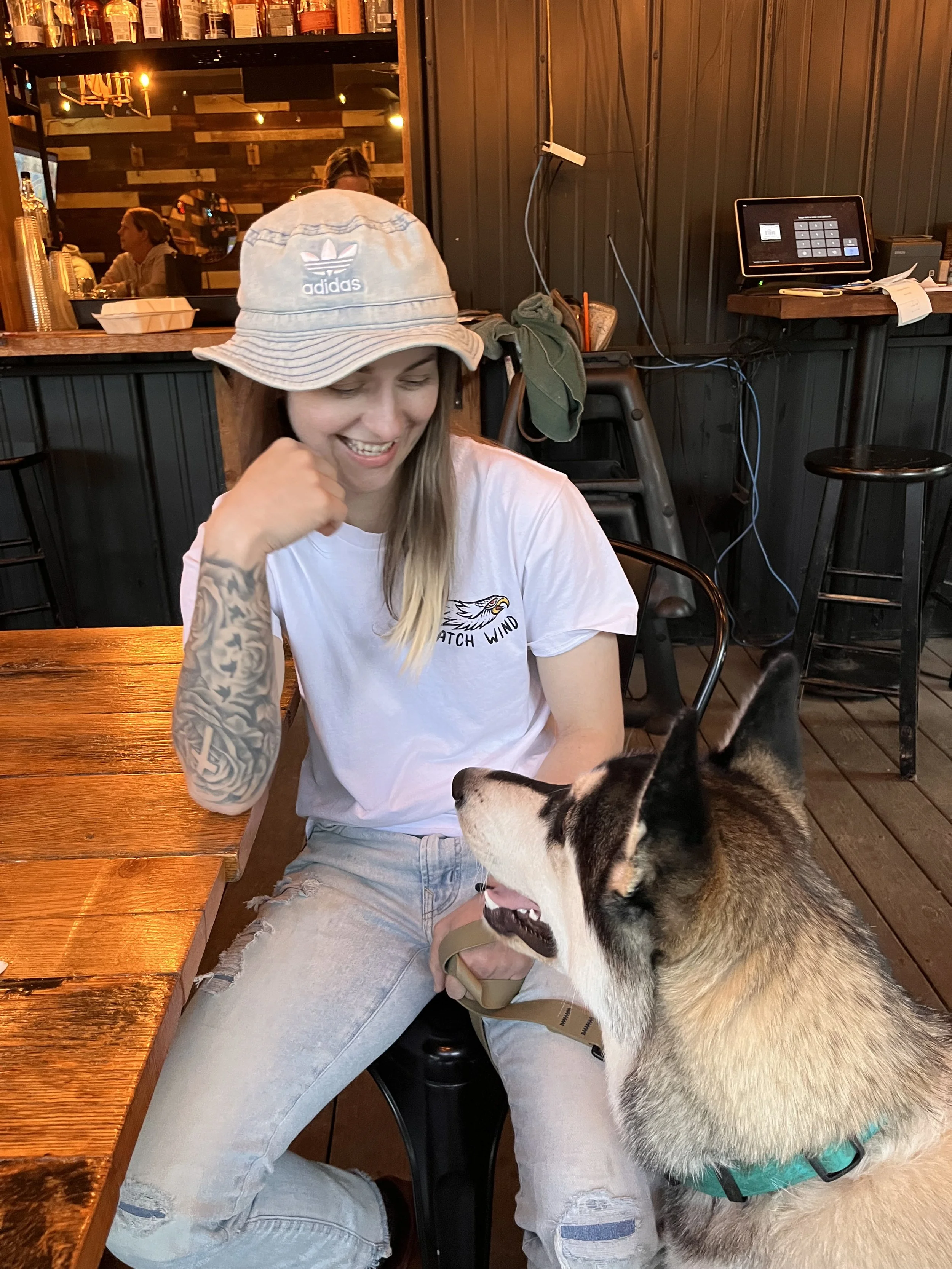 A woman with a tattooed arm wearing a white Adidas hat and a white t-shirt sitting at a wooden table in a restaurant, smiling at a large Husky dog sitting in front of her.