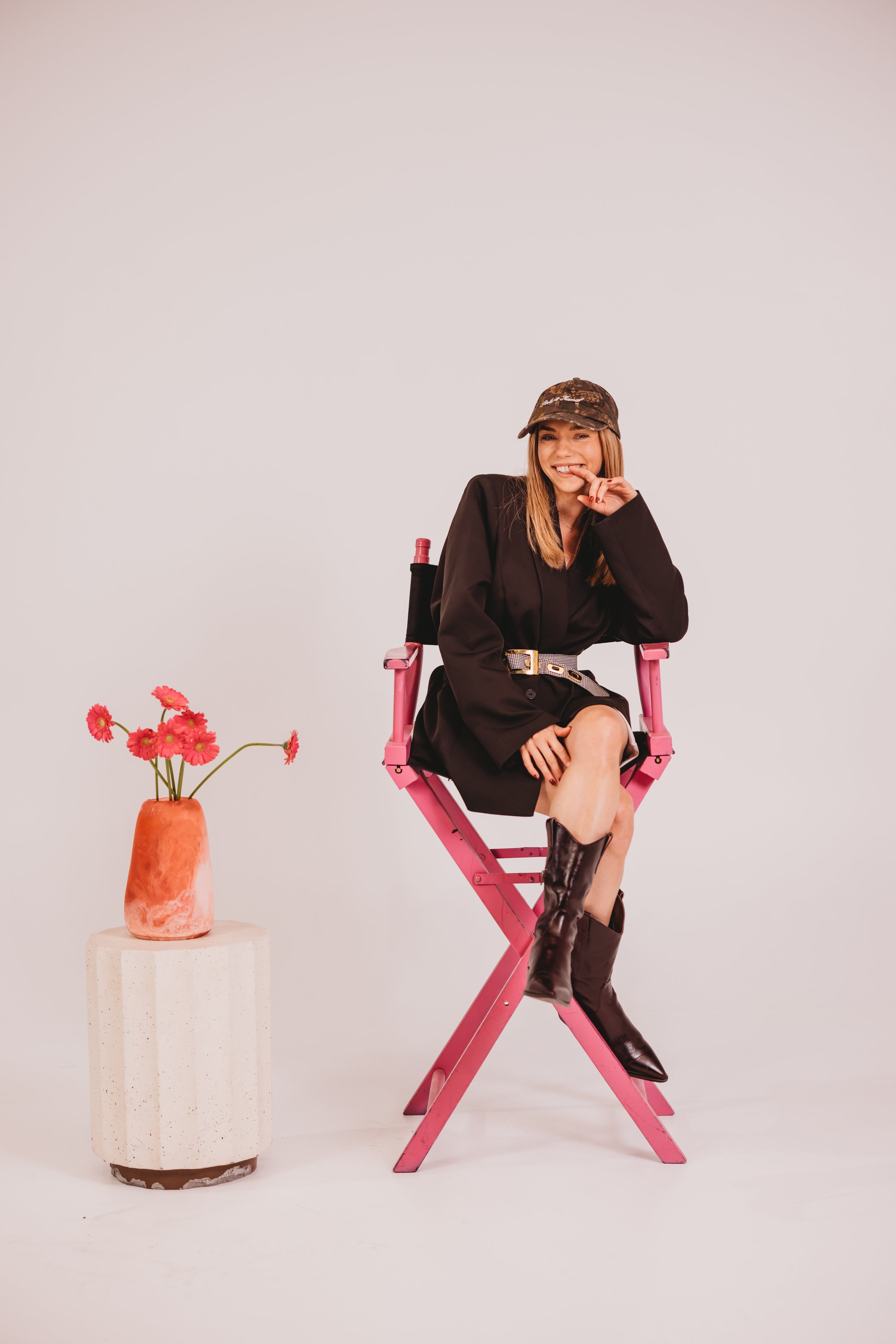 A young woman sitting on a pink director's chair, wearing a black dress, black boots, and a camouflage cap, smiling and making a playful gesture with her hand near her face, next to a white pedestal with a pink flower vase containing pink flowers.